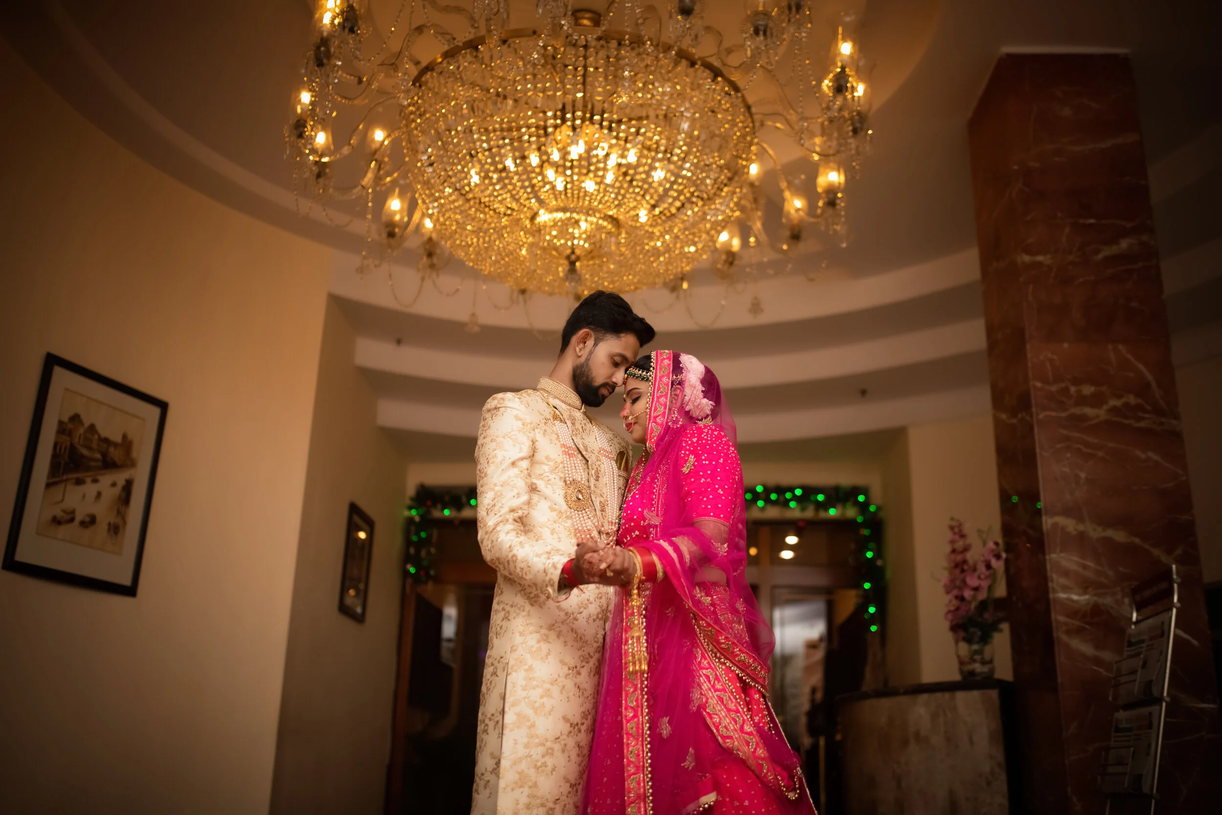 An Indian bride and groom in traditional attire share a dance underneath a large chandelier, indoors with decorative lights and artwork on the walls.