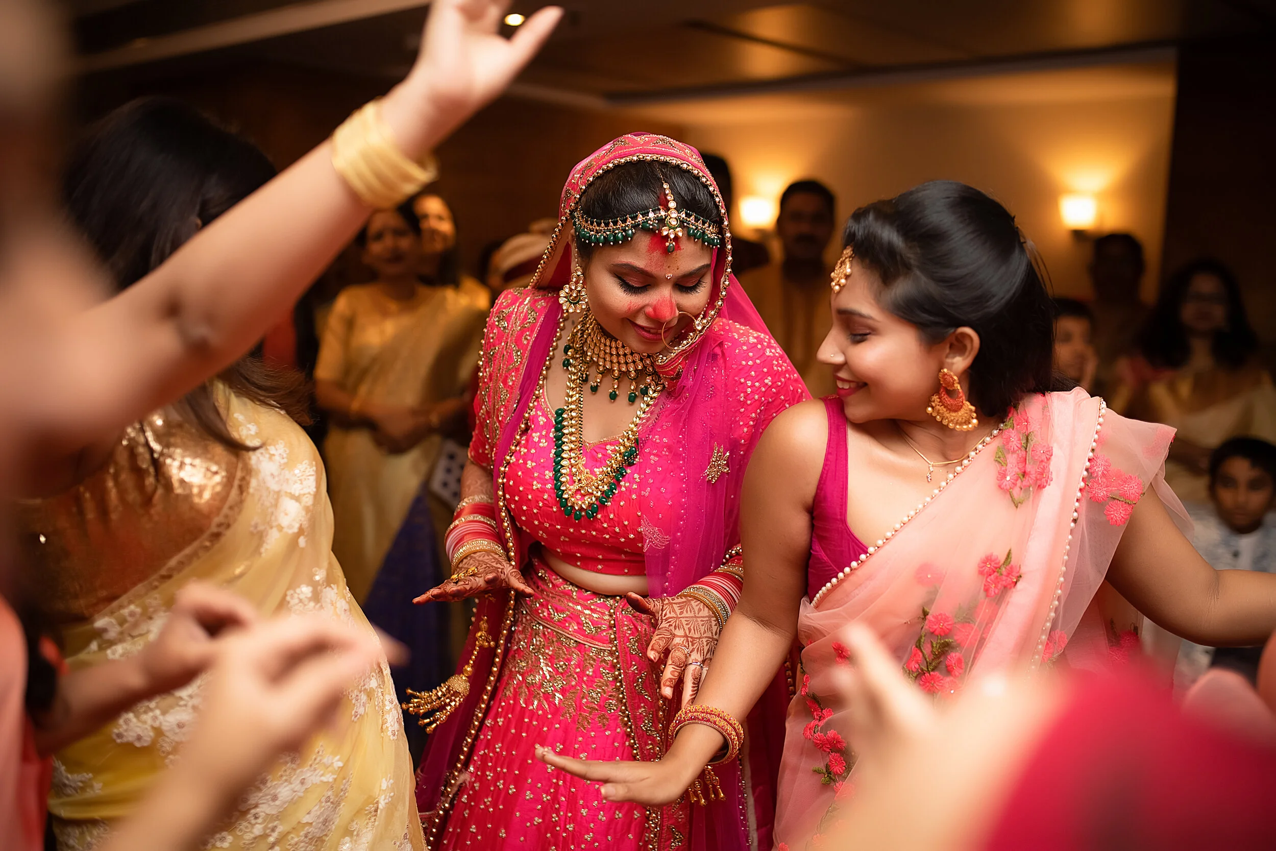 Indian women dancing at a celebration, with the bride in a pink traditional outfit and jewelry, and another woman in a pink saree smiling.