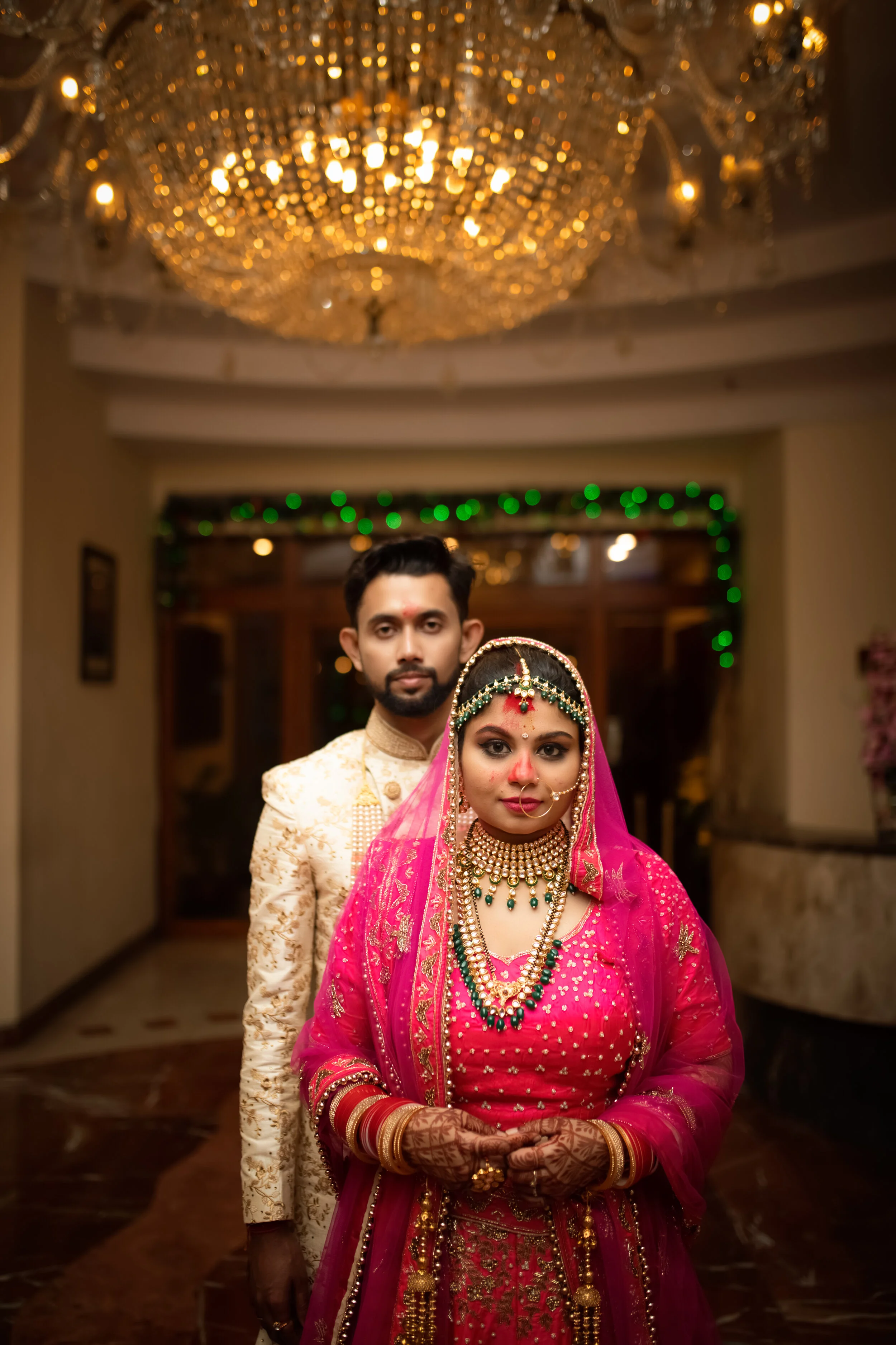 A bride and groom in traditional Indian wedding attire standing indoors, with a grand chandelier overhead and festive green lighting in the background.