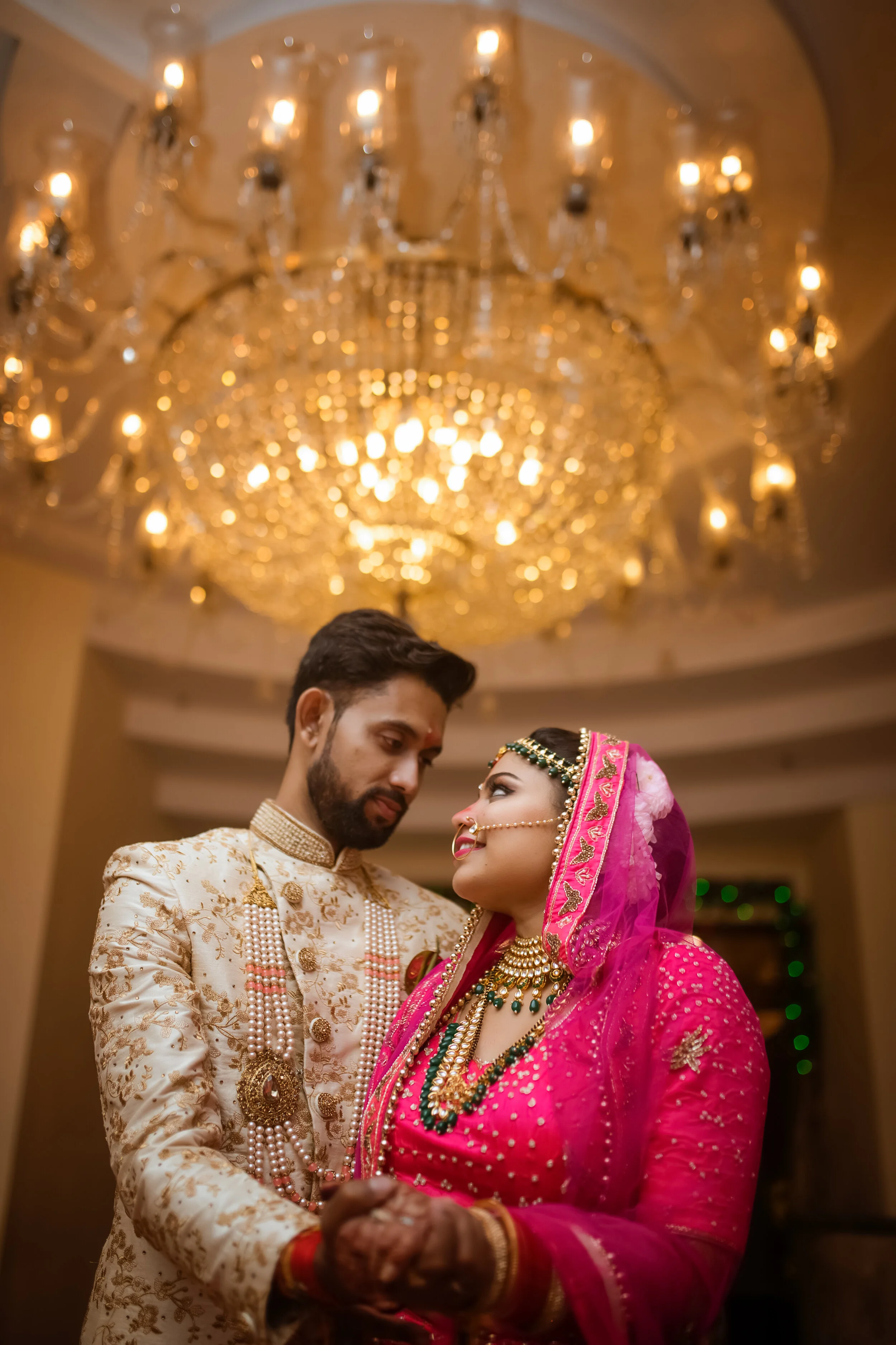 Indian bride and groom dressed in traditional wedding attire, holding hands and looking at each other under a grand chandelier.