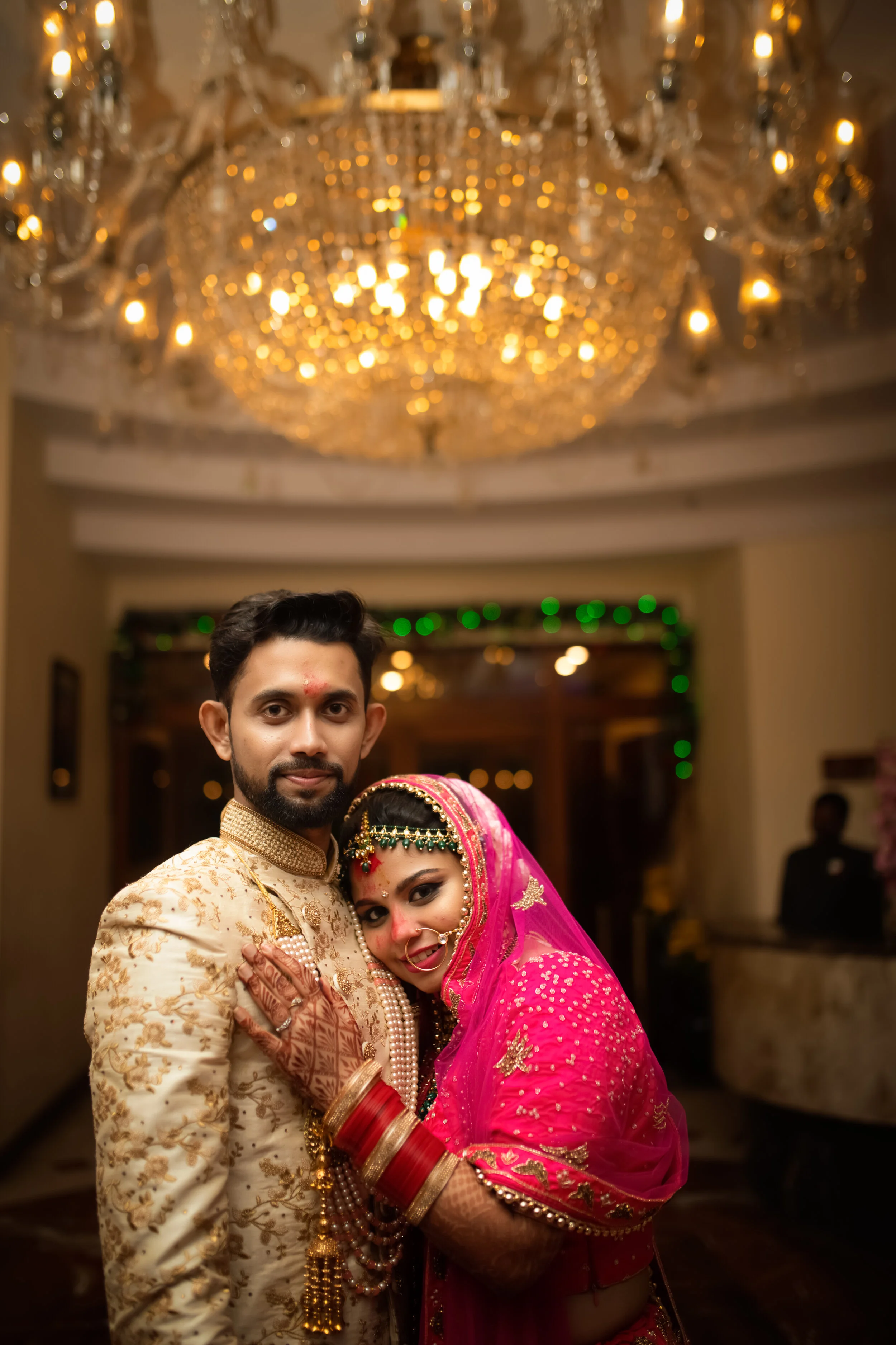 A couple dressed in traditional Indian wedding attire at their wedding celebration, with a grand chandelier above and warm lighting in the background.