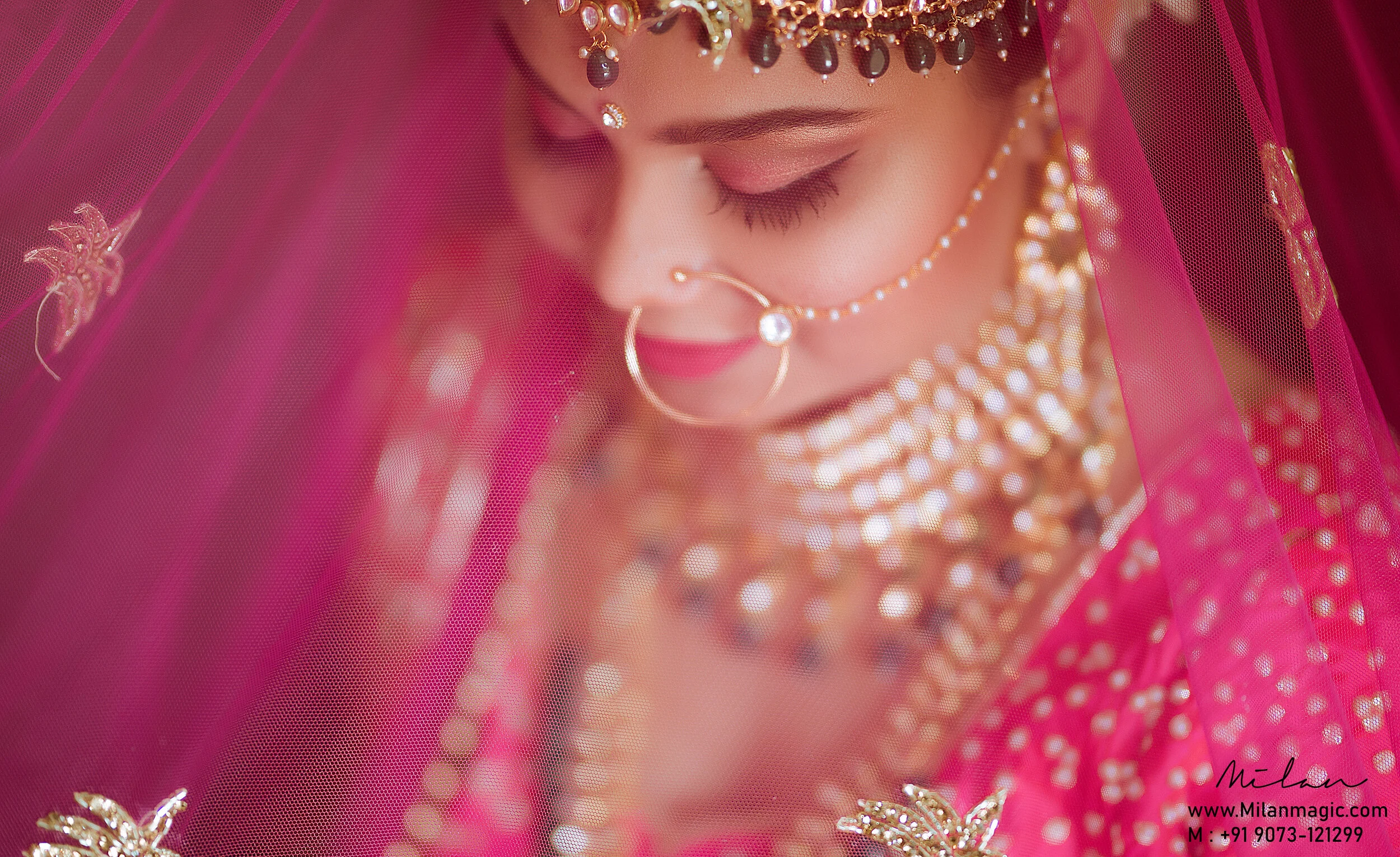 A woman dressed in traditional Indian bridal attire with a pink veil covering her face, adorned with gold jewelry including a nose ring, a necklace, and earrings, with her eyes closed.