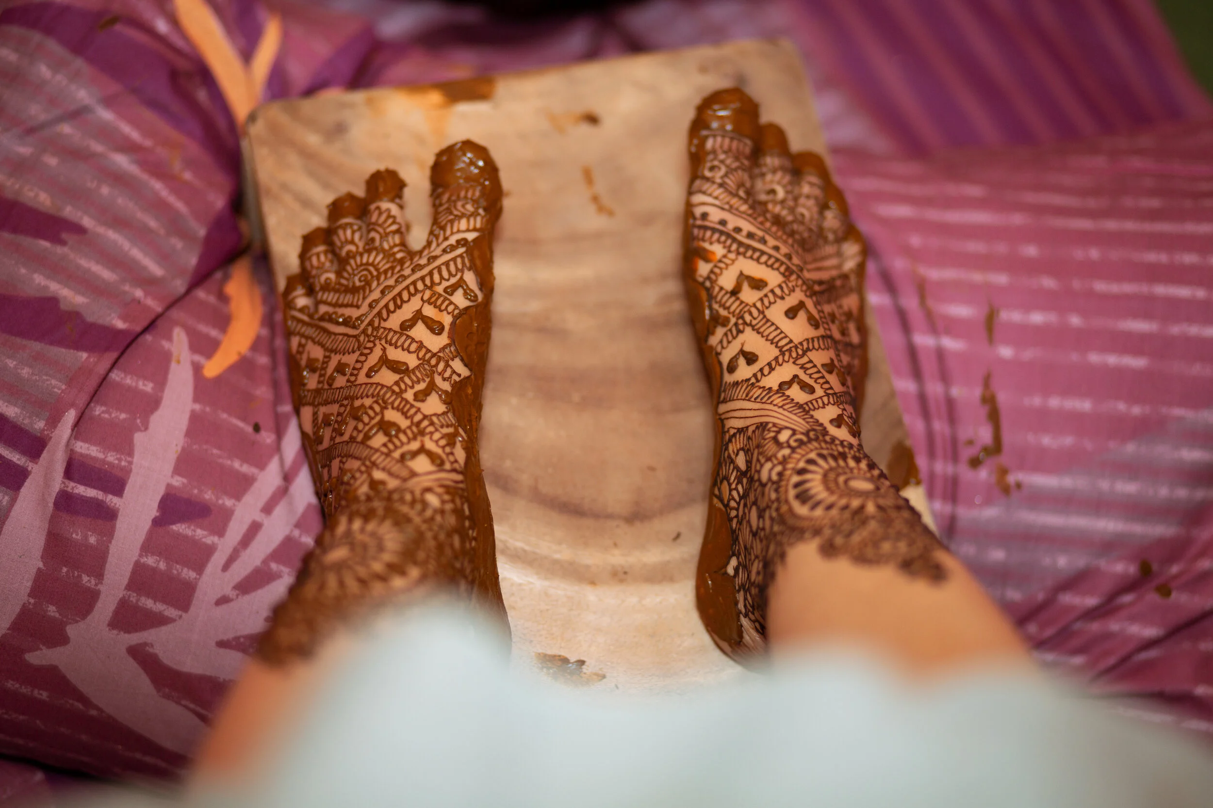 Hands with intricate henna designs applying henna on a surface, with a pink patterned fabric in the background.