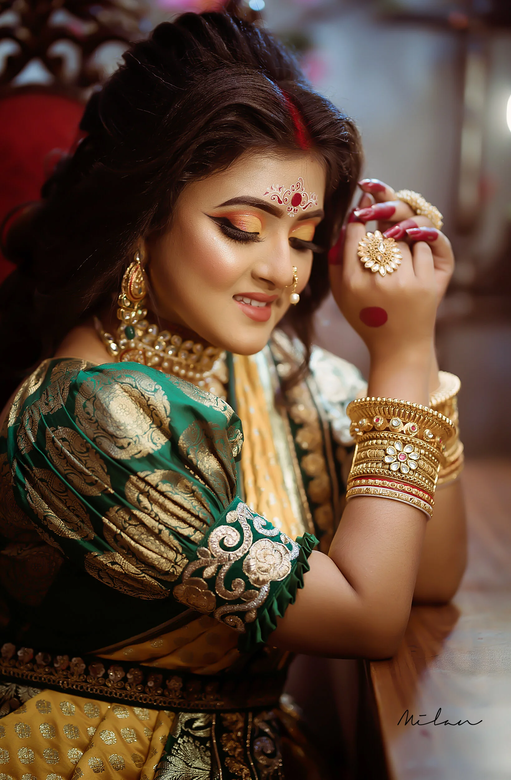 A woman dressed in traditional Indian attire, wearing ornate jewelry, with her eyes closed, smiling gently, and resting her head on her arm. She has jewelry on her nose, ears, neck, and wrists, and vibrant makeup with orange and gold eyeshadow.
