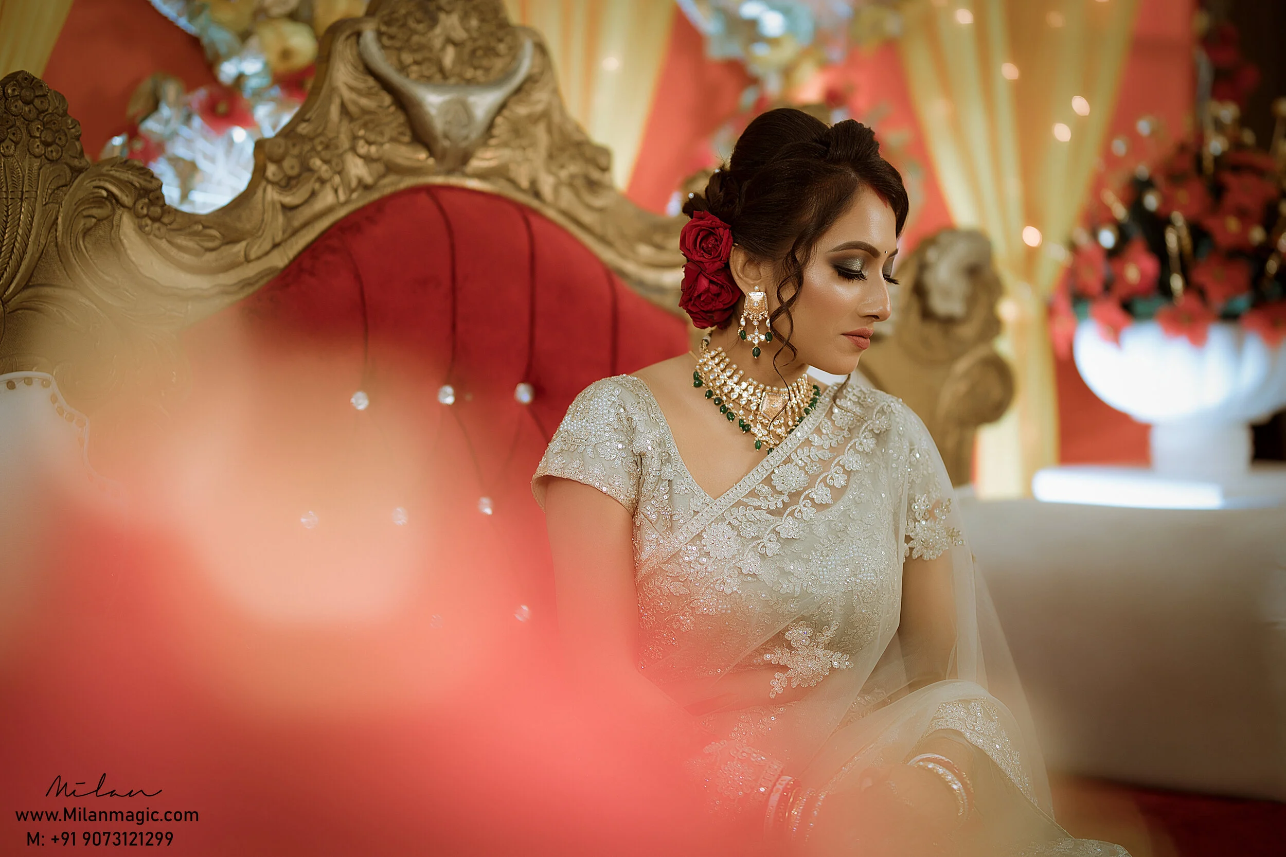 A woman dressed in traditional Indian bridal attire sitting on an ornate red and gold throne with decorative background, wearing jewelry and red roses in her hair.