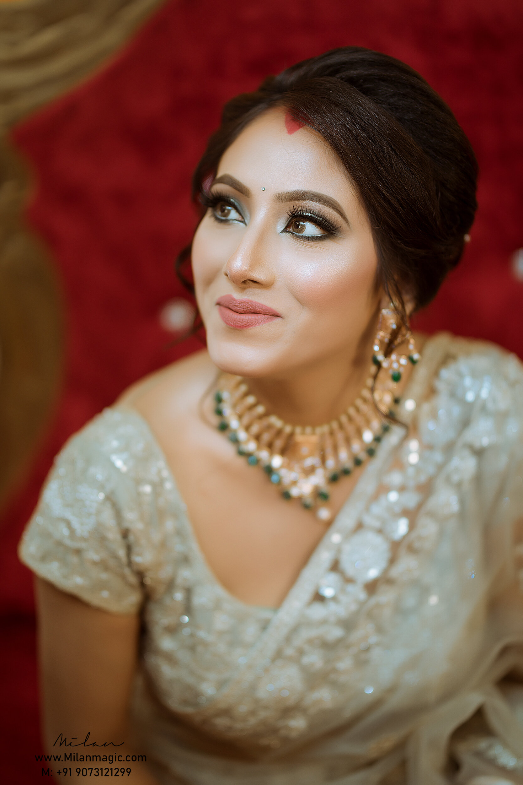 Close-up of a woman with makeup and jewelry, wearing an embellished beige outfit, posing against a red background.