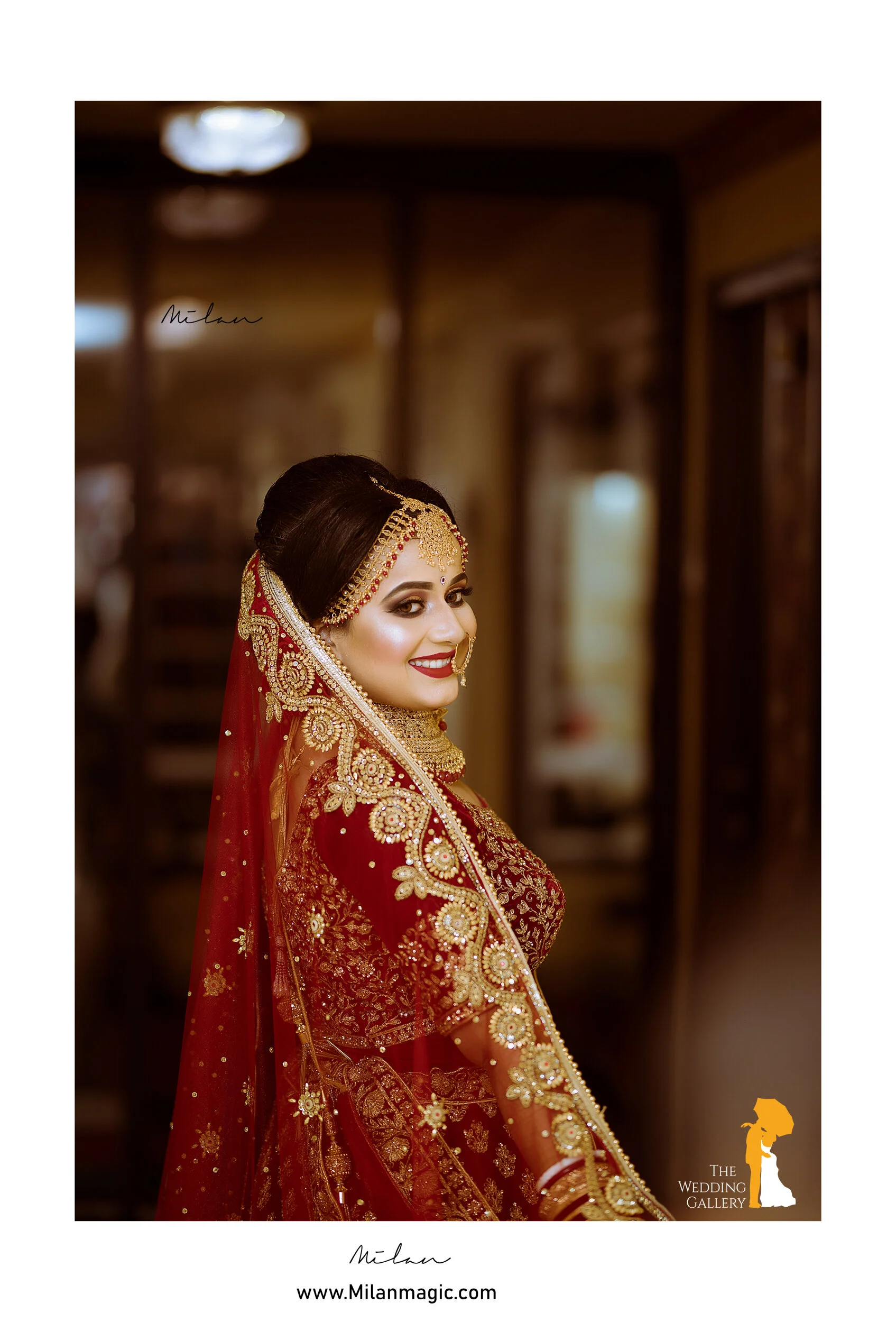 A bride dressed in traditional red and gold bridal attire with detailed embroidery, wearing gold jewelry and a veil, smiling and looking over her shoulder indoors.