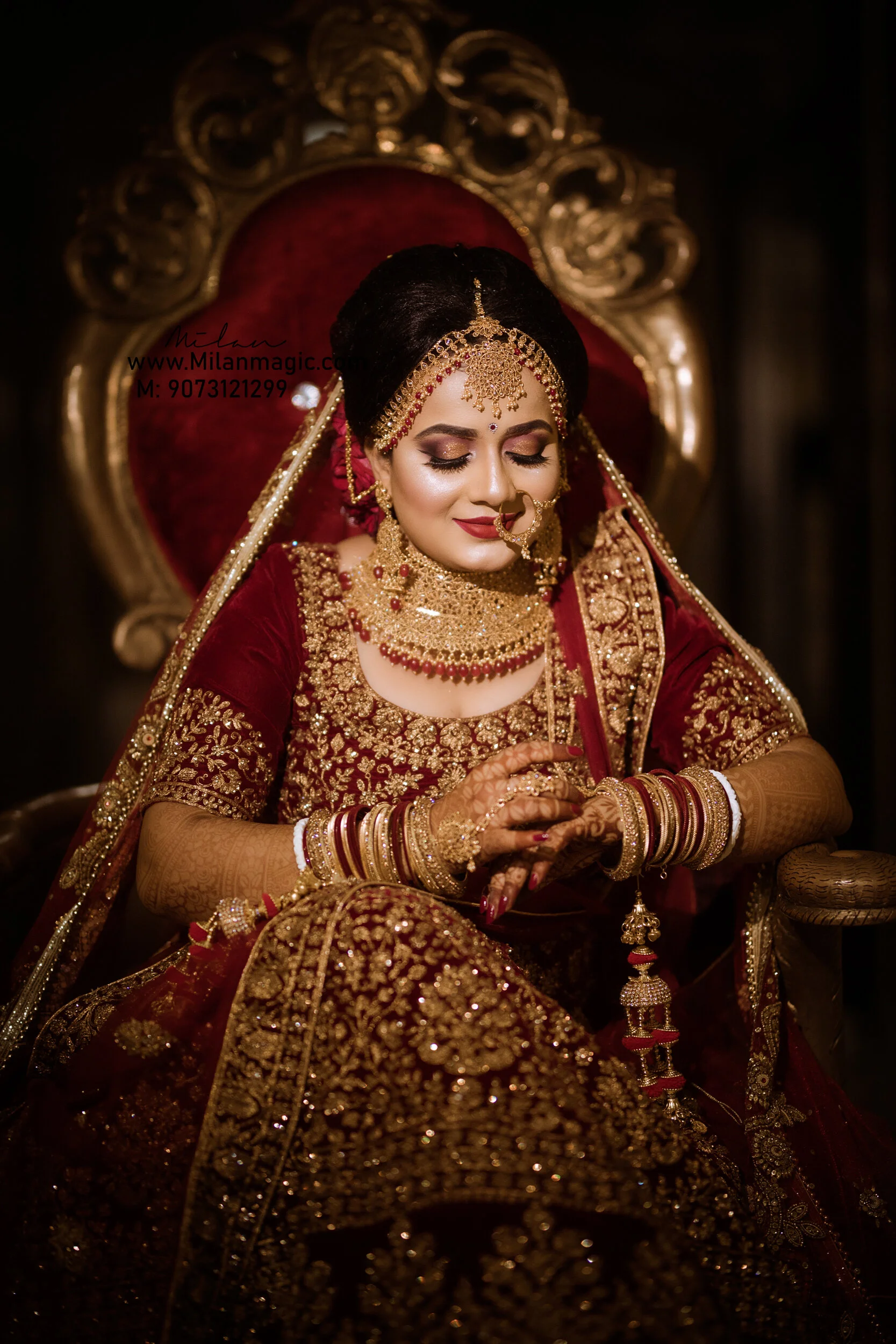 A bride dressed in traditional Indian wedding attire, sitting on an ornate chair, wearing a deep red and gold embroidered outfit with matching jewelry, including a necklace, earrings, nose ring, and headpiece.
