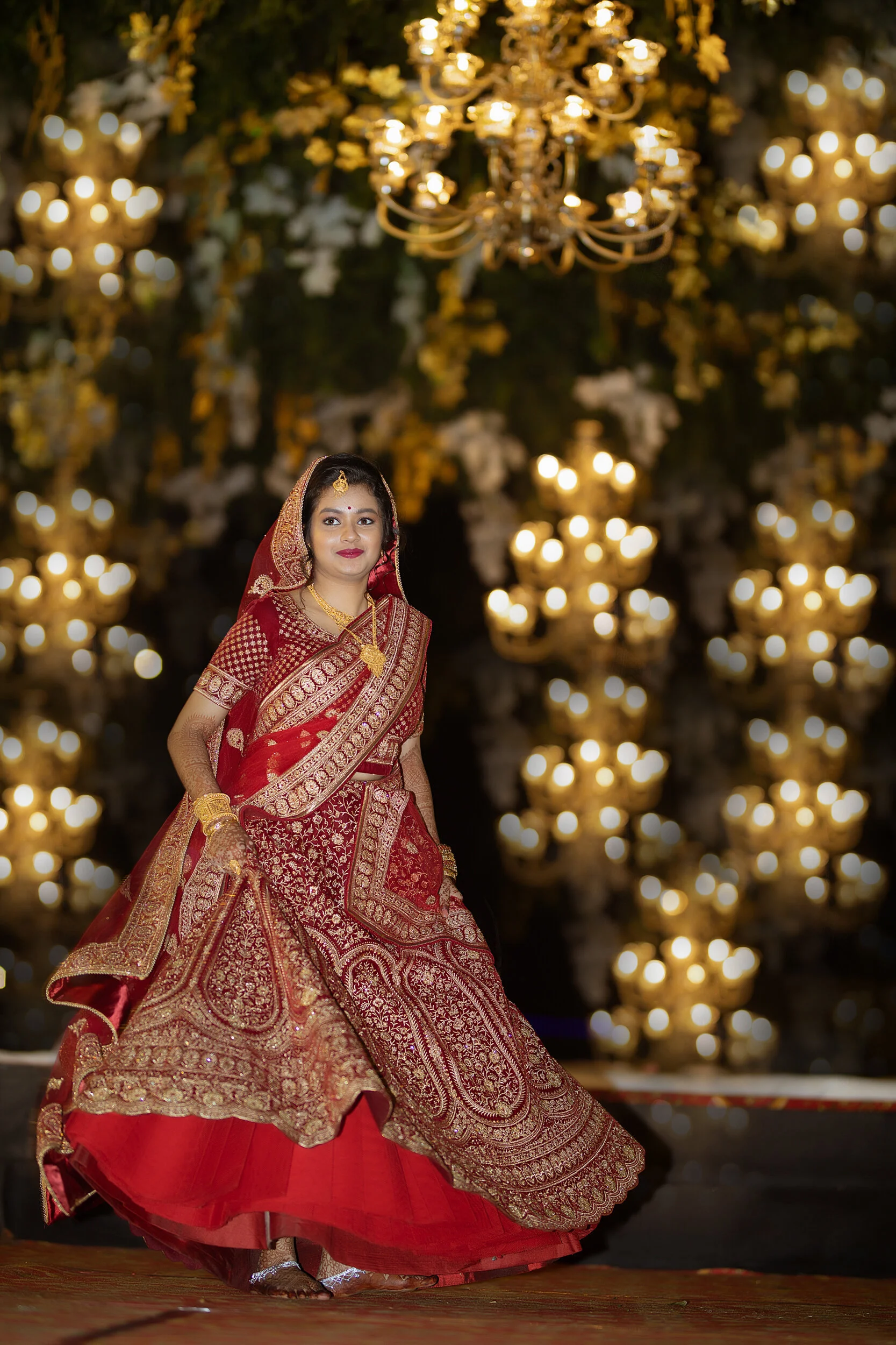 A woman dressed in a traditional red and gold Indian bridal outfit, standing in front of a backdrop of large, ornate chandeliers and decorated with flowers, during a wedding celebration.