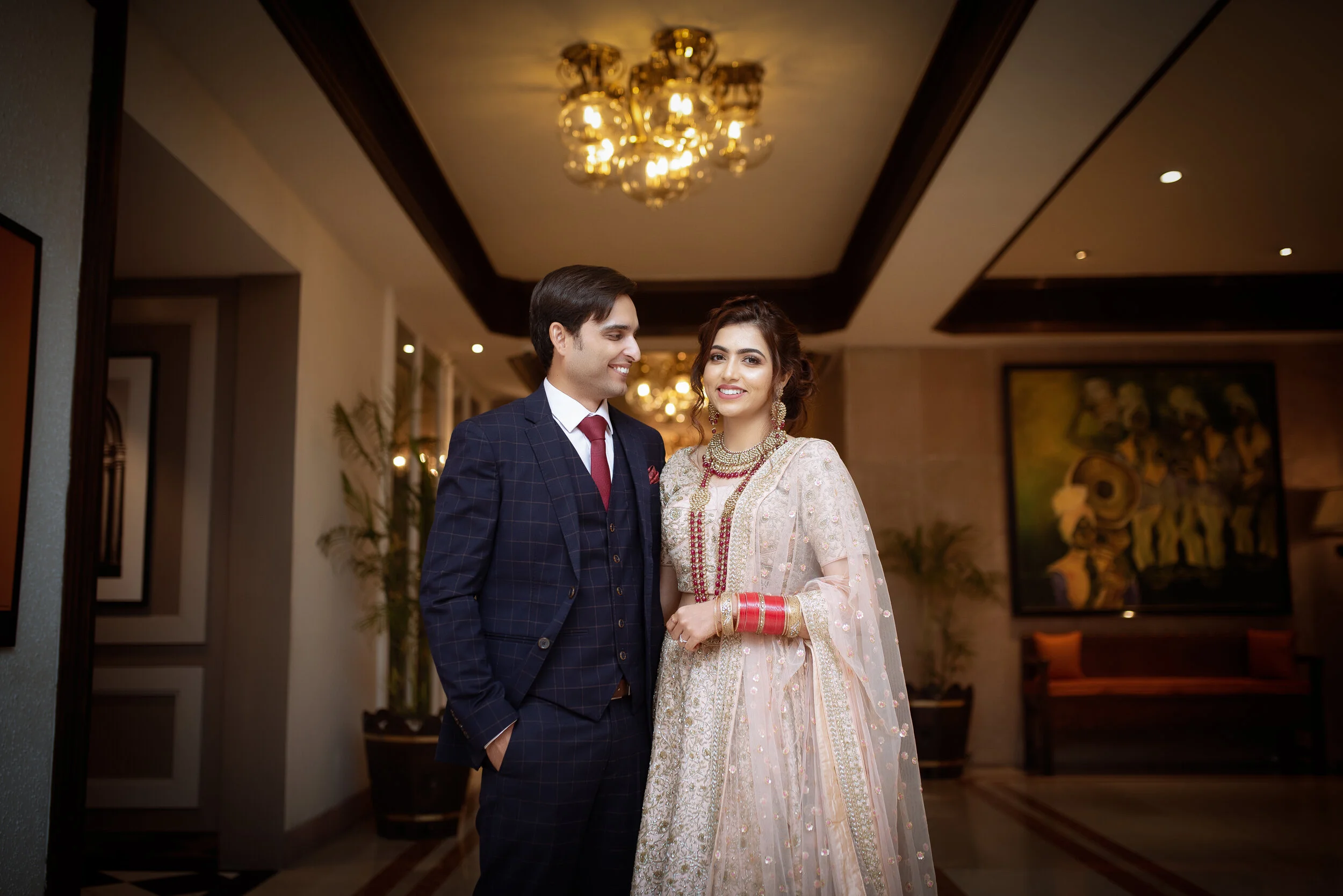 A man and woman dressed in traditional Indian wedding attire, standing inside a well-lit hotel lobby, smiling.