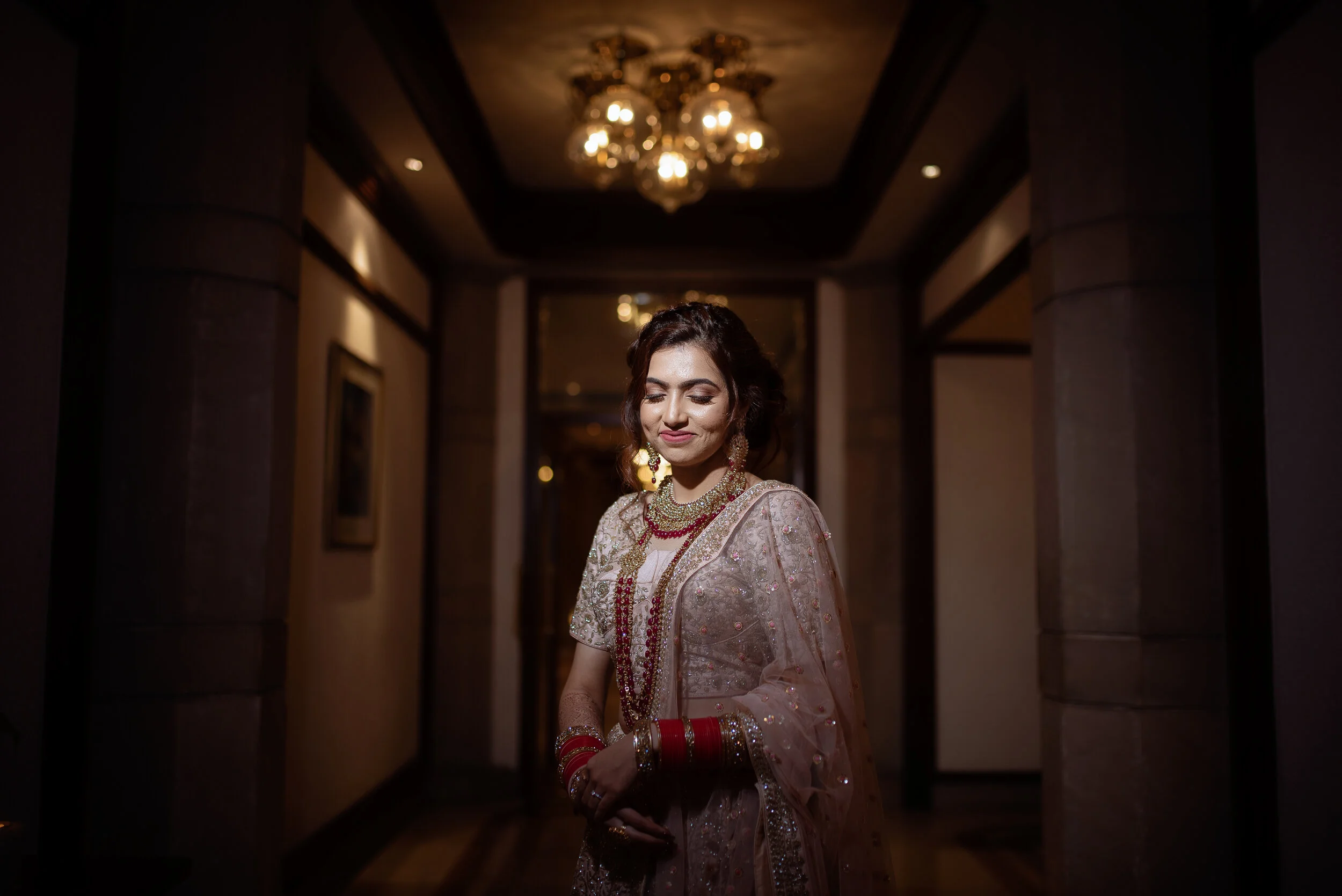 A woman dressed in traditional Indian attire standing in a dimly lit hallway with a chandelier overhead. She is wearing a detailed, embellished outfit with gold jewelry, red bangles, and earrings, with her eyes closed and a slight smile.