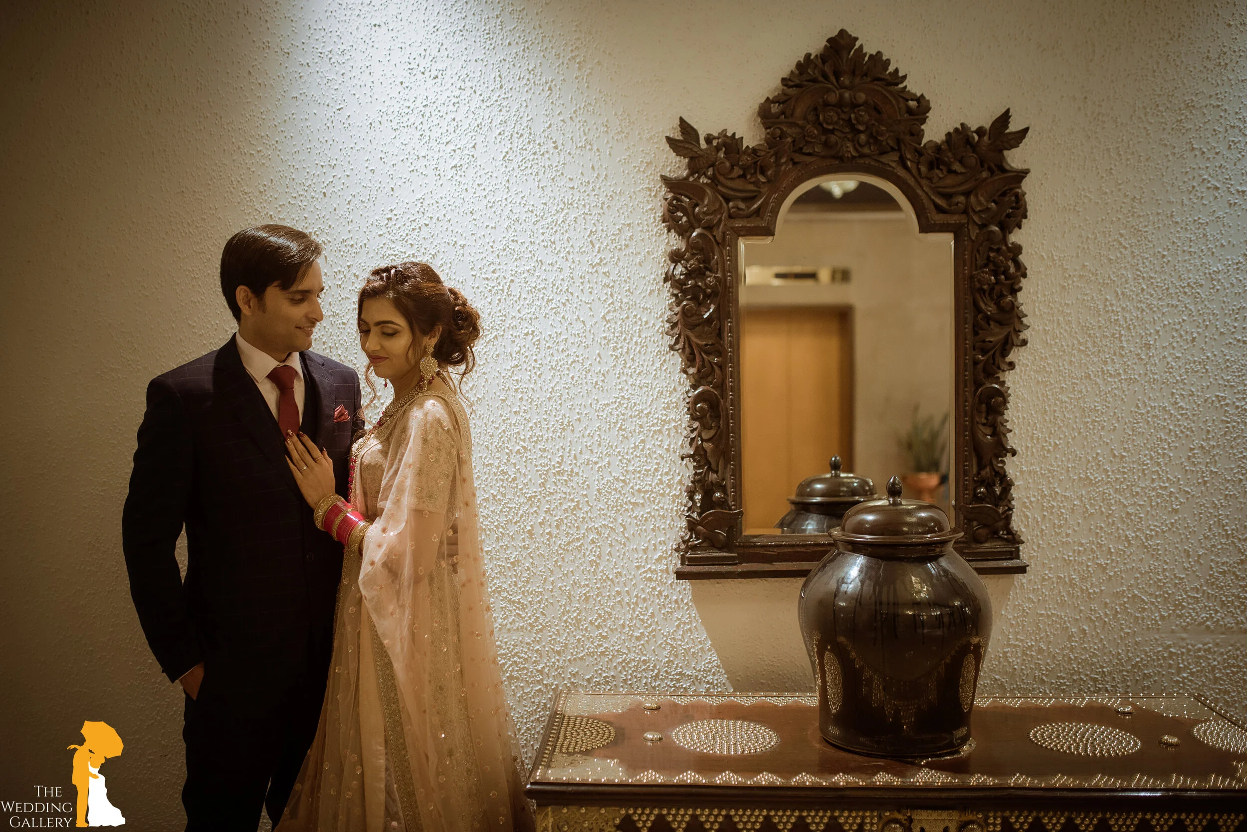 A bride and groom standing close together indoors, with the bride dressed in traditional Indian attire and the groom in a dark suit, reflected in a decorative mirror hanging on a textured wall, beside a wooden table with large ceramic jars.