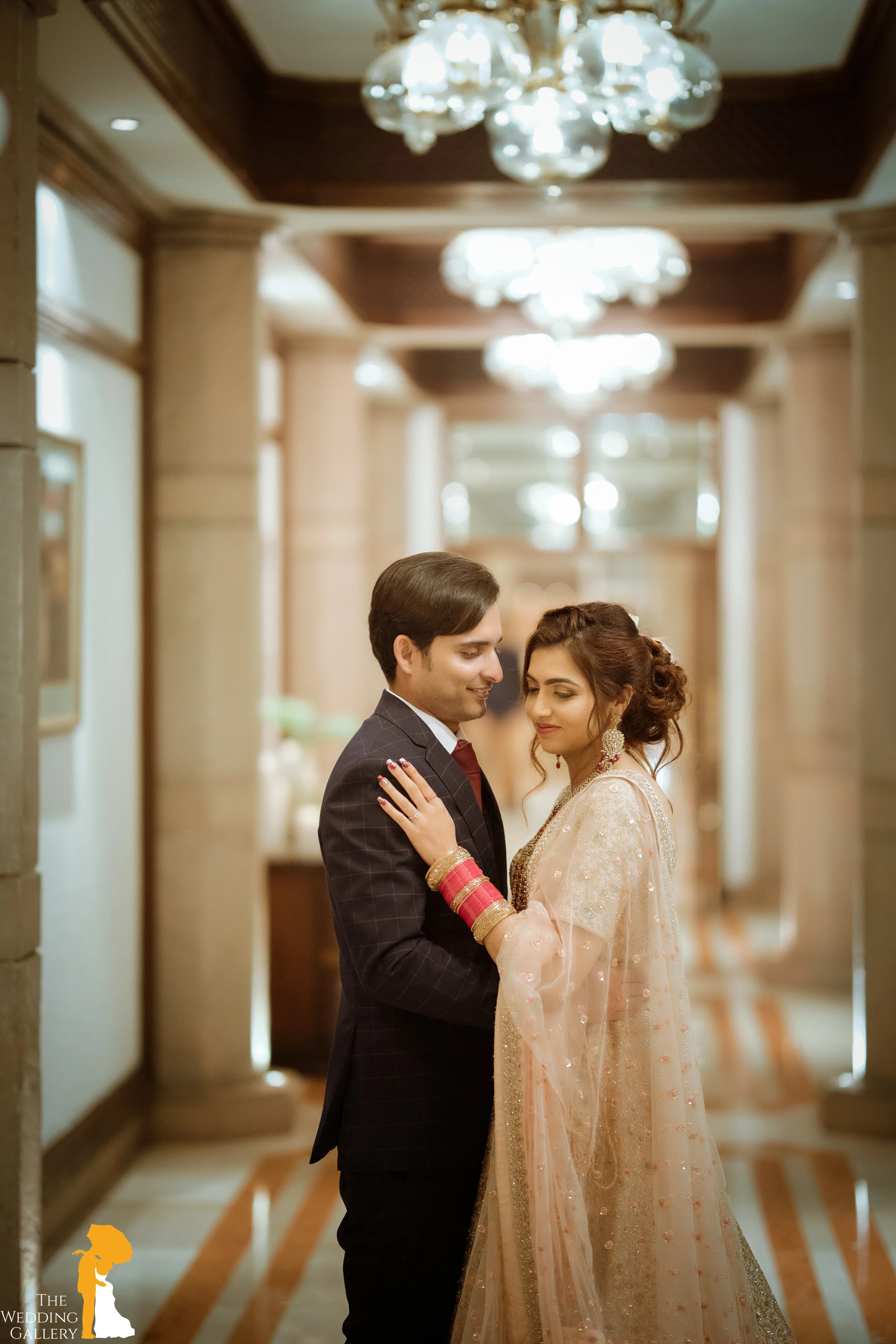 A bride and groom sharing a tender moment in an elegant, well-lit hallway. The bride is wearing a light pink, embellished traditional dress and red bangles, while the groom is in a dark plaid suit. They are holding each other affectionately.
