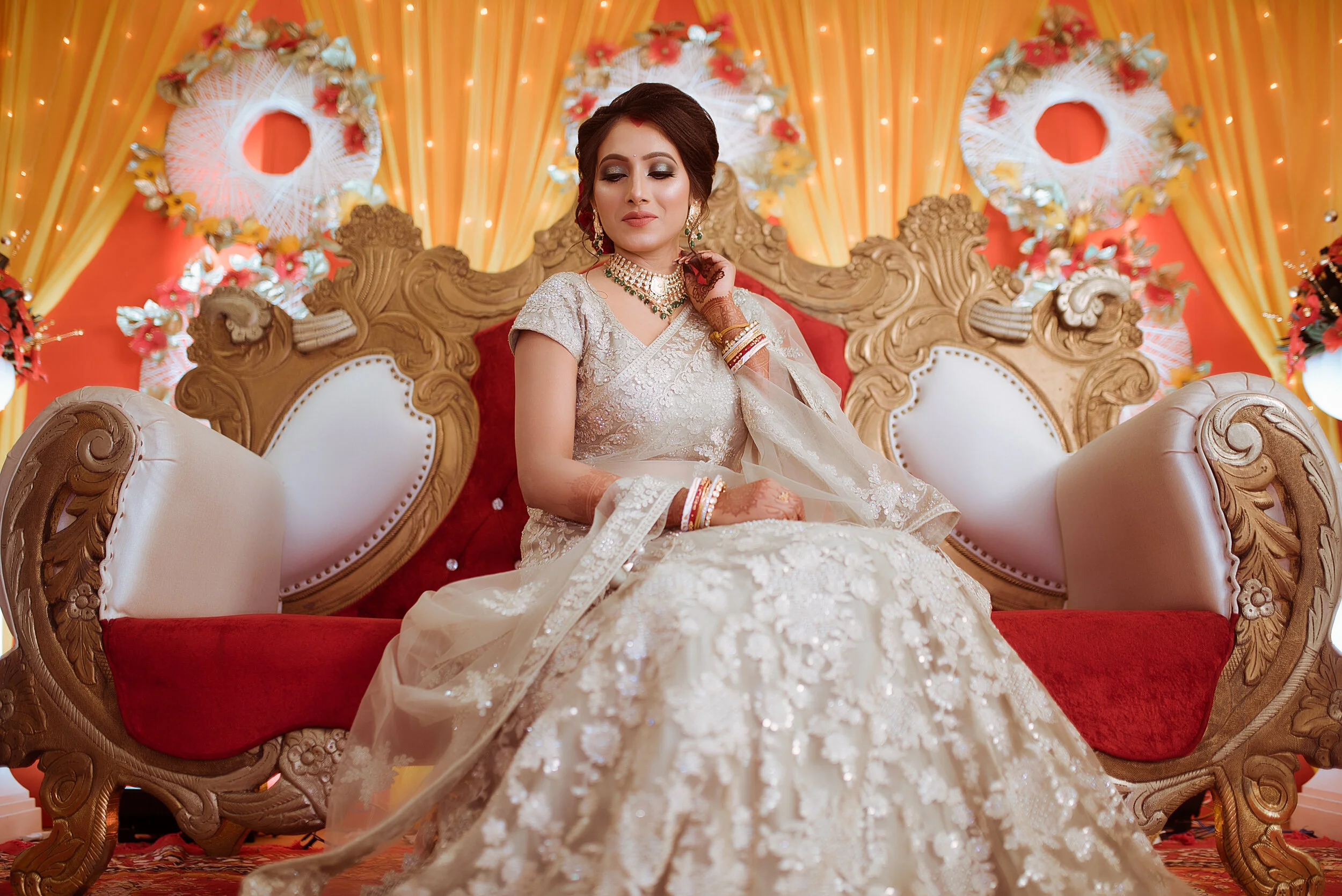 A woman dressed in a cream-colored wedding gown with embroidery and lace detail, sitting on an ornate wooden and white bench with red upholstery, surrounded by orange and red drapery and floral decorations, wearing jewelry and bangles.