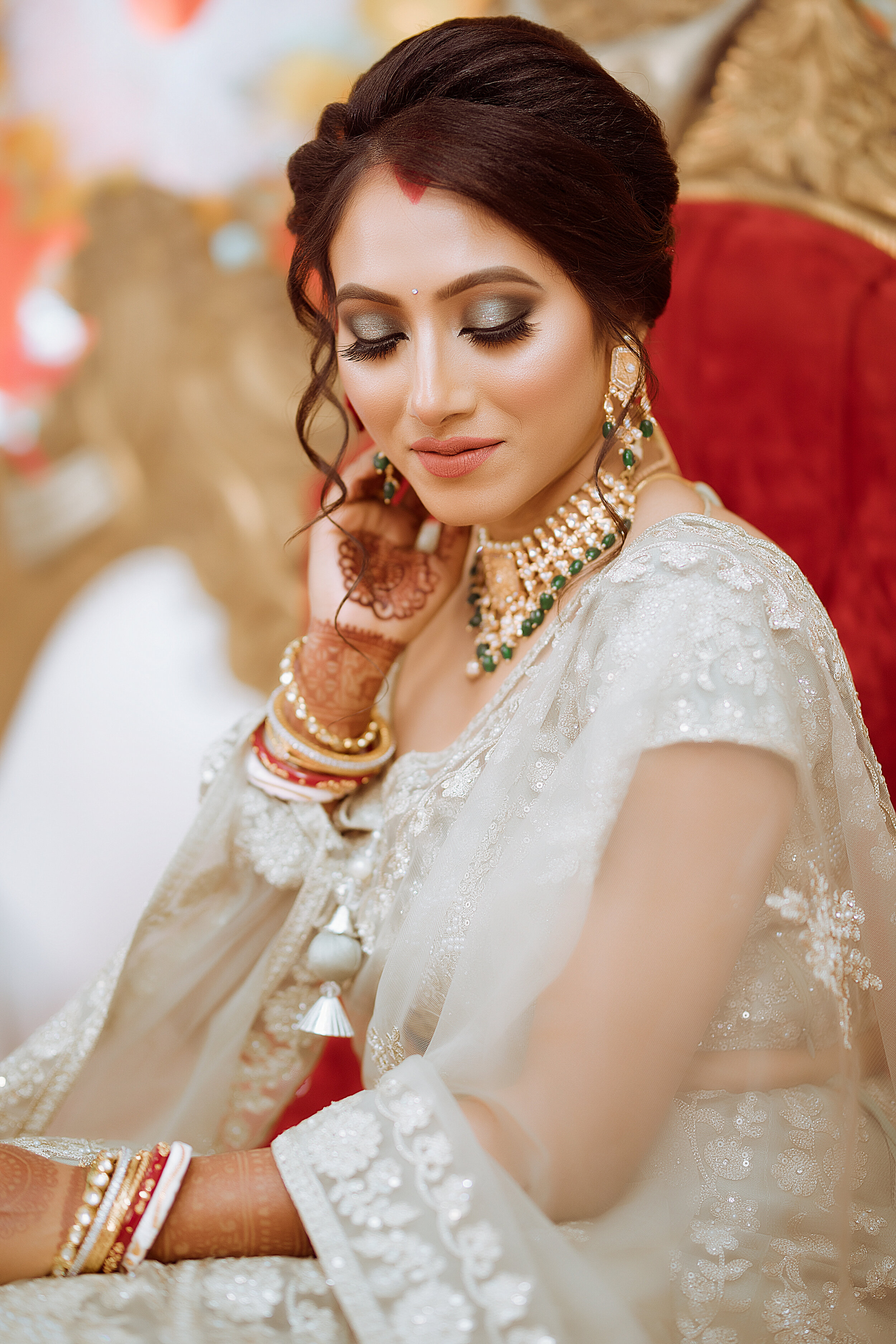 A bride with closed eyes, wearing traditional Indian attire with jewelry and mehndi on her hand, sitting on a red and gold chair.