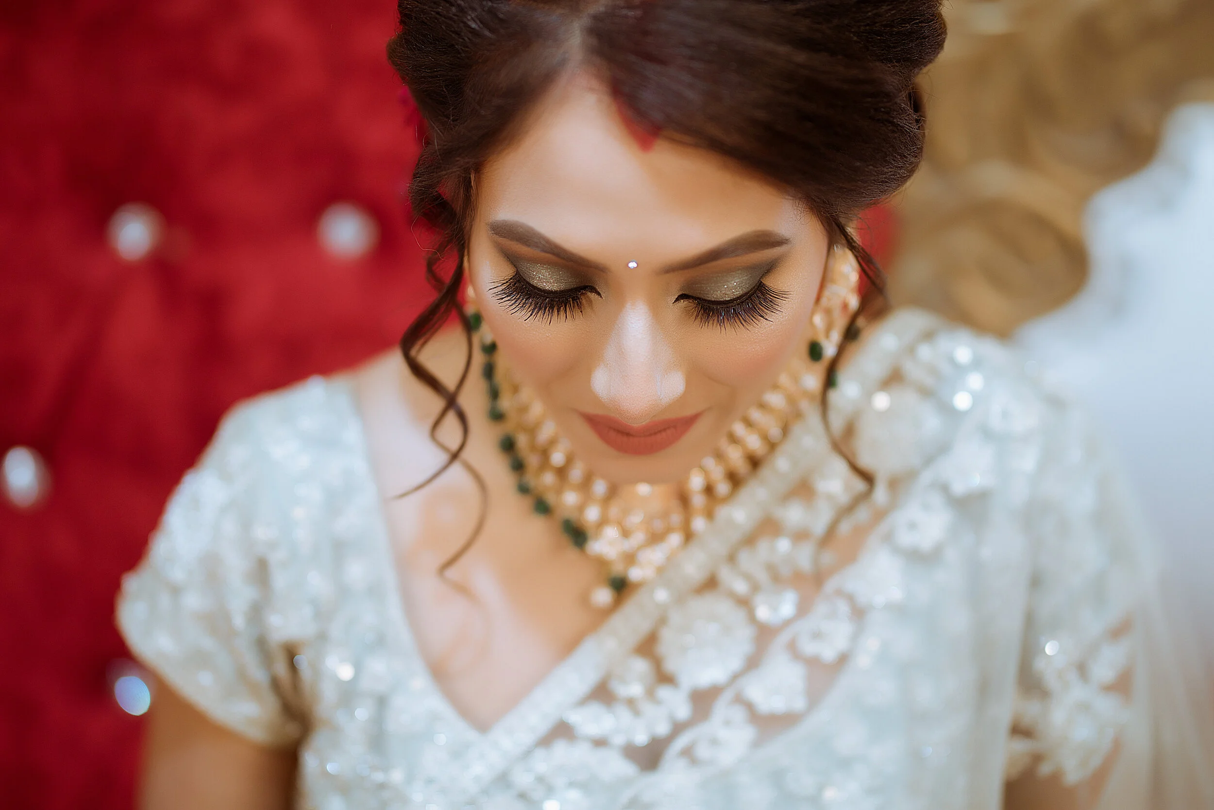 A woman dressed in traditional Indian wedding attire, with elaborate makeup and jewelry, resting her head on a red, tufted chair with pearl embellishments.