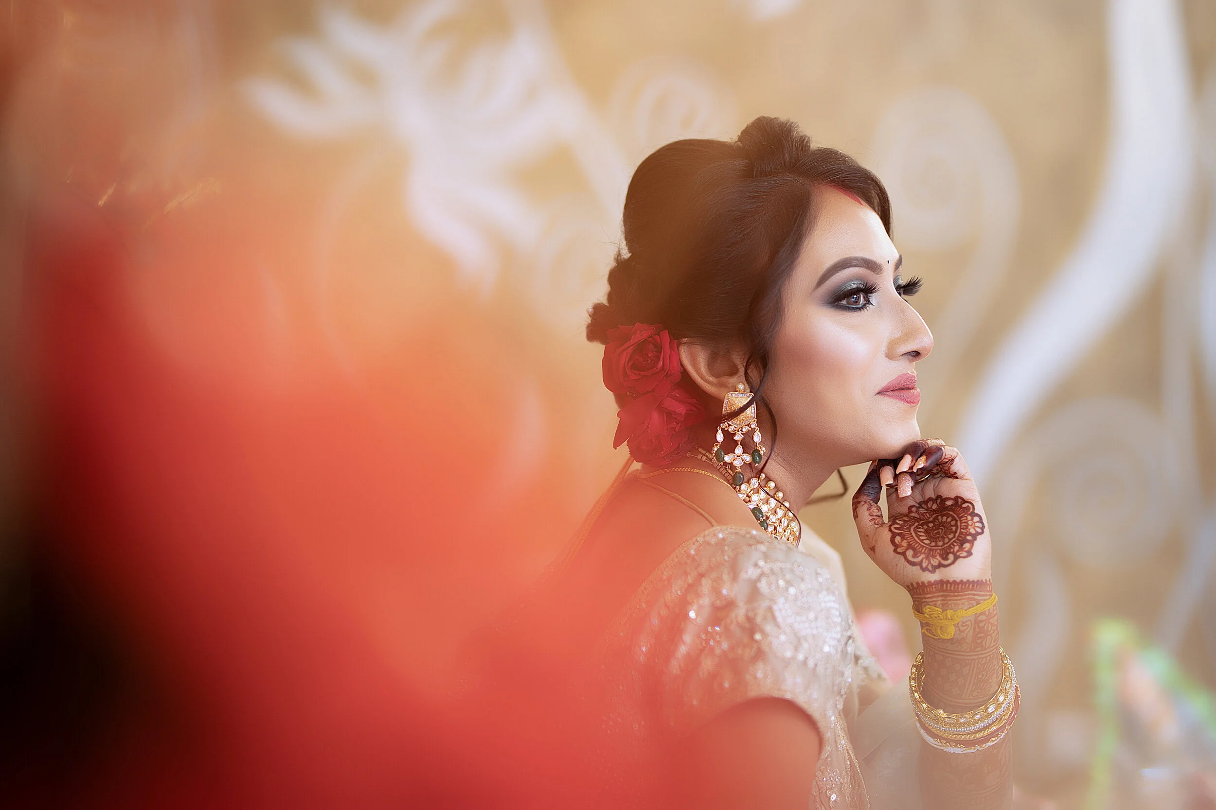 A woman dressed in traditional Indian attire, adorned with jewelry and flowers in her hair, gazing thoughtfully to the side with a blurred, decorative background.