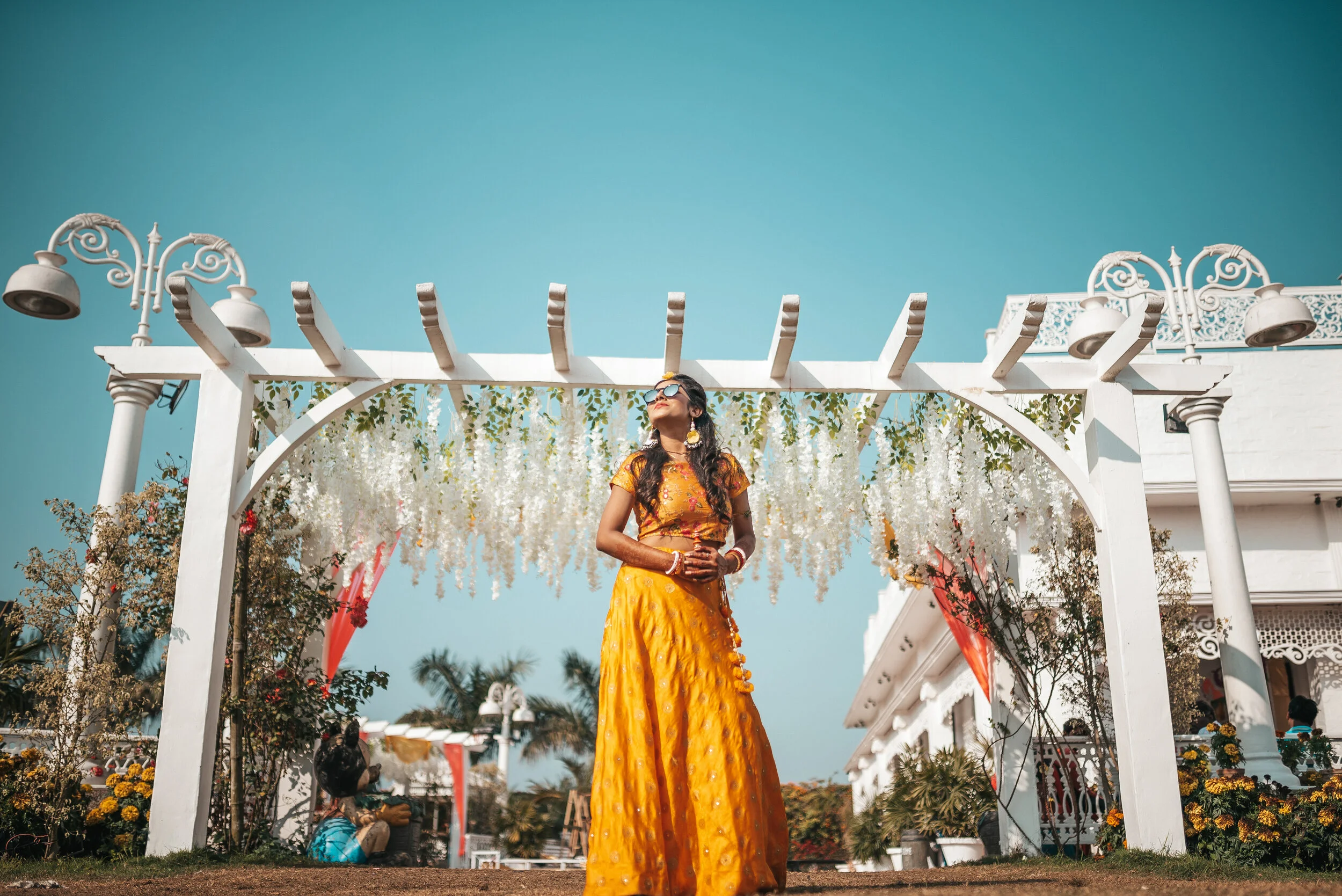 A woman in a yellow traditional dress stands under a white decorative arch with hanging white flowers, outdoors with clear blue sky and palm trees in the background.