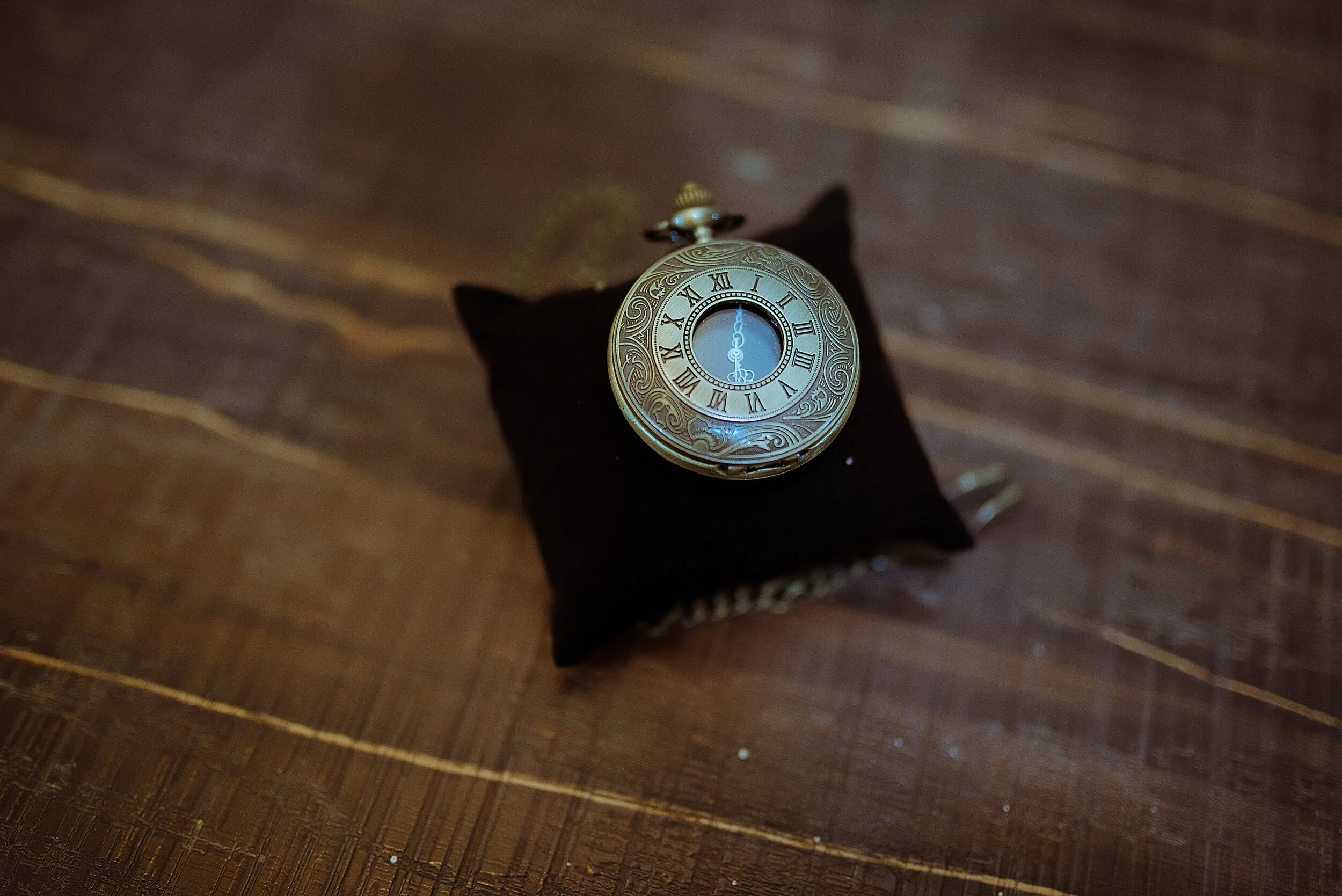 A vintage pocket watch with Roman numerals on the face, placed on a small black fabric cushion on a wooden surface.