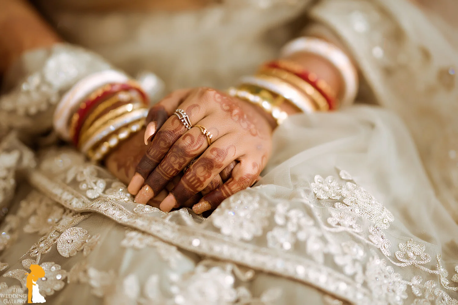 Close-up of hands resting on embroidered fabric, adorned with henna designs, gold rings, bangles, and jewelry, likely at a wedding or festive occasion.