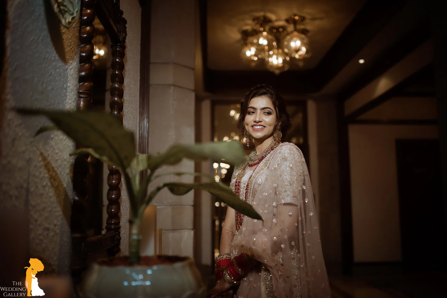 A woman dressed in traditional Indian attire, smiling, standing indoors in front of a mirror and a decorative wall, wearing jewelry and red bangles, with a plant in the foreground.