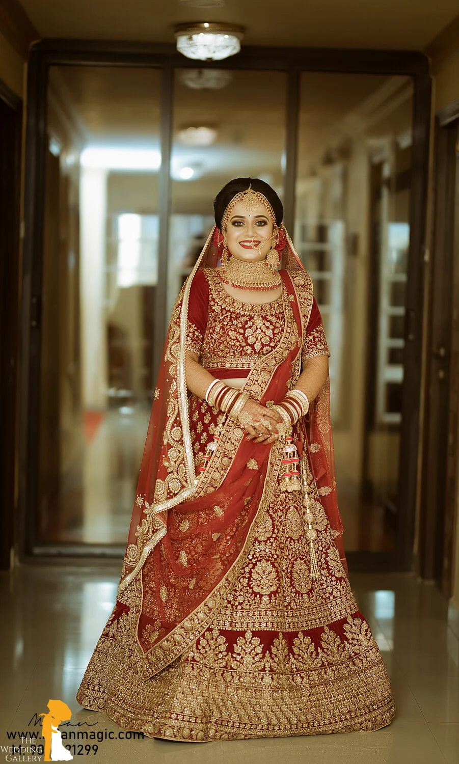 A bride standing indoors dressed in a red and gold heavily embroidered traditional wedding outfit, adorned with jewelry, bangles, and a head covering, smiling at the camera.