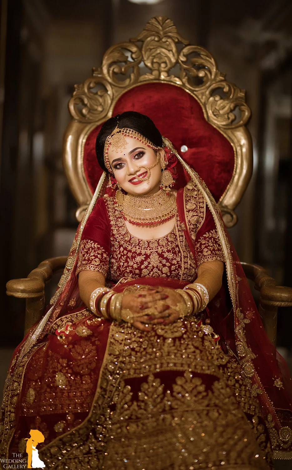 A bride in elaborate red and gold traditional wedding attire, sitting on a large ornate chair with a red velvet backrest, smiling happily.
