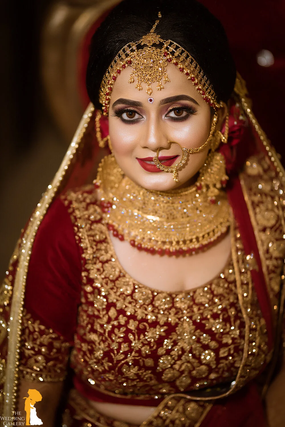 A woman dressed in traditional Indian bridal attire with gold jewelry, red and gold embroidery, and elaborate makeup.