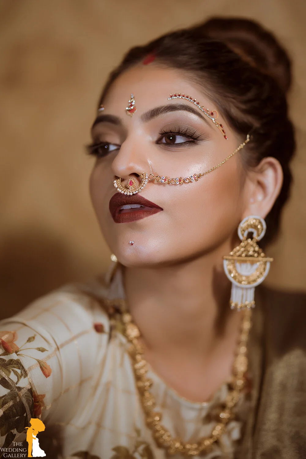 Close-up of a woman wearing traditional Indian jewelry and makeup, with a decorative nose ring connected to her ear by a chain, large earrings, and intricate forehead adornments, dressed in a cream-colored outfit with floral embroidery.