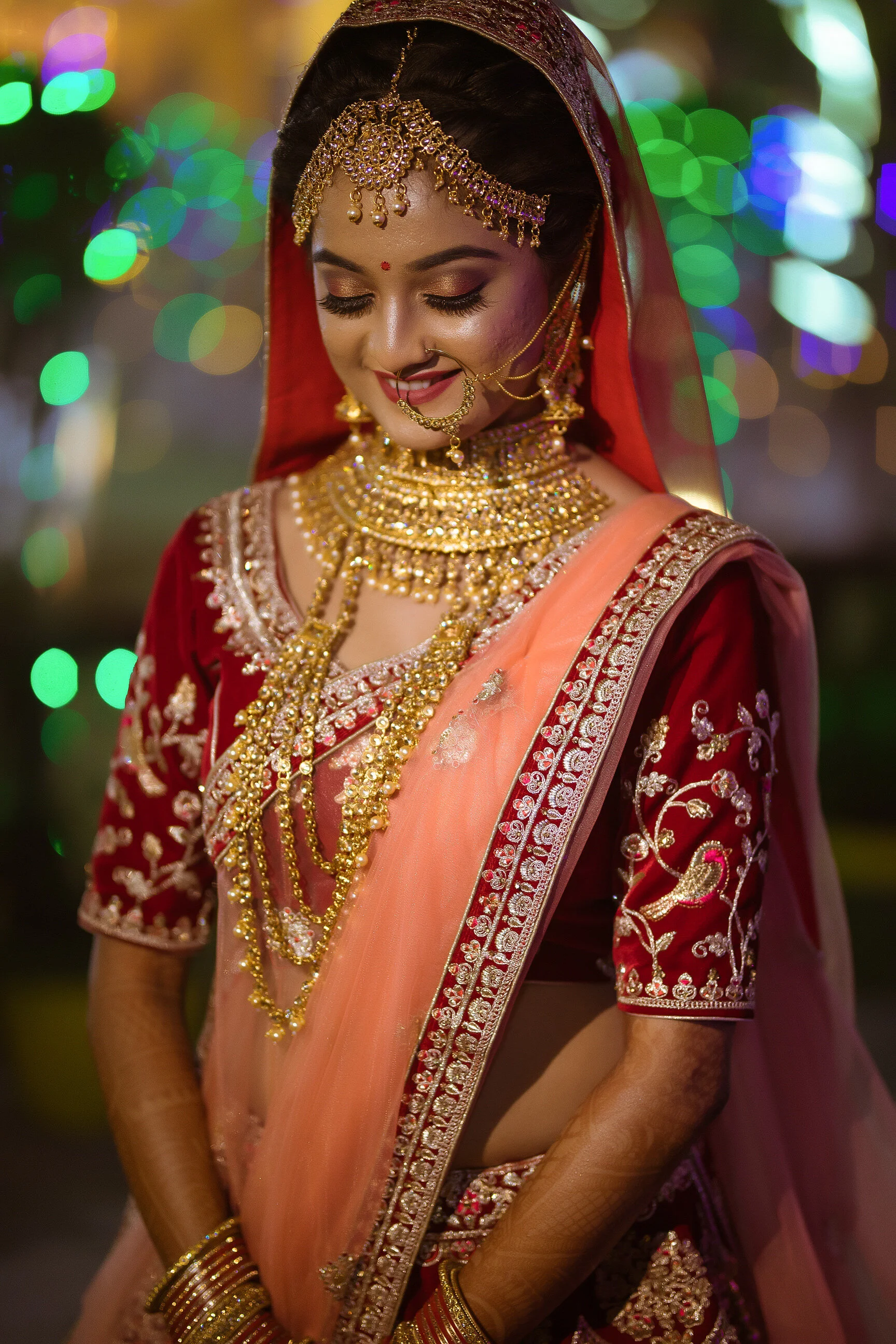 A woman dressed in traditional Indian bridal attire, wearing red and gold embroidered outfit with a peach dupatta. She is adorned with elaborate gold jewelry including a choker necklace, layered necklaces, earrings, maang tikka, nose ring, and bangle