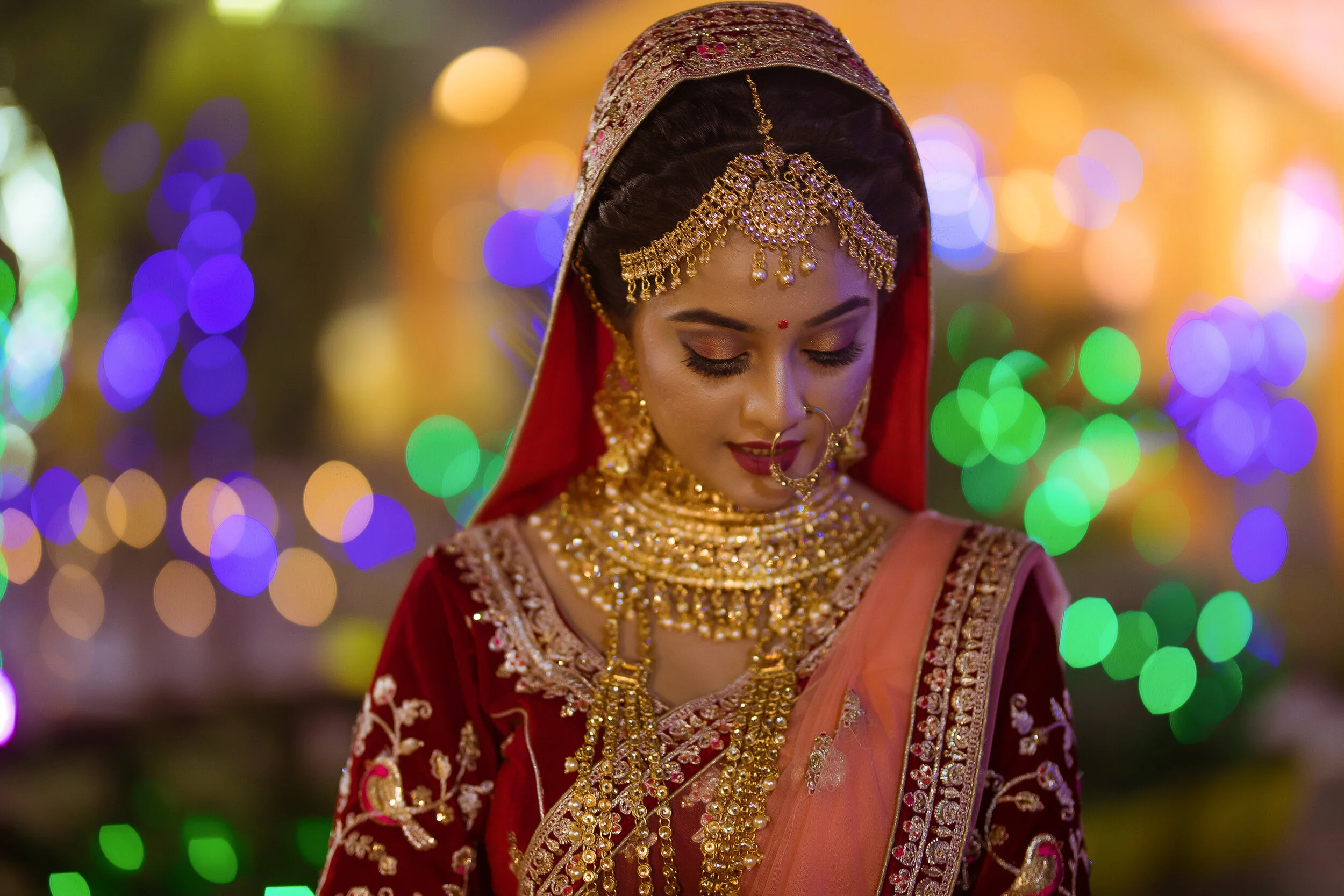 A woman dressed in traditional Indian wedding attire with intricate gold jewelry, red and orange saree, and a decorative headpiece, standing with her eyes closed against a backdrop of colorful blurred lights.
