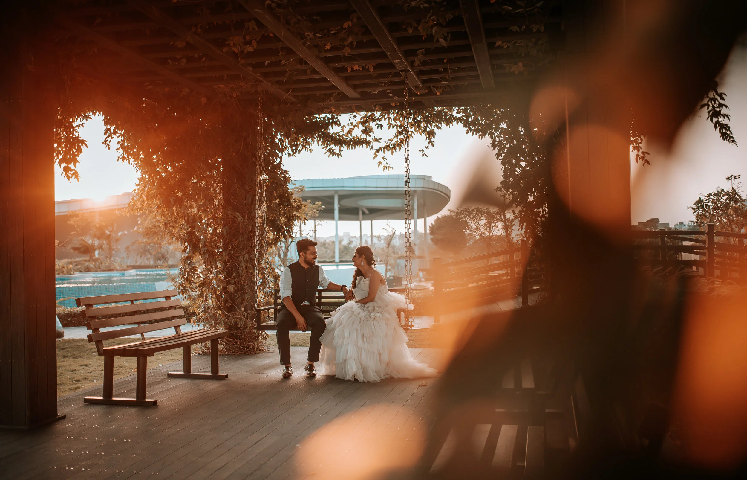 A couple sitting on a swing bench under a wooden pergola with vines, having a conversation during sunset. The woman wears a white wedding dress, and the man wears a dark vest and white shirt. The background shows a pool and a modern outdoor structure