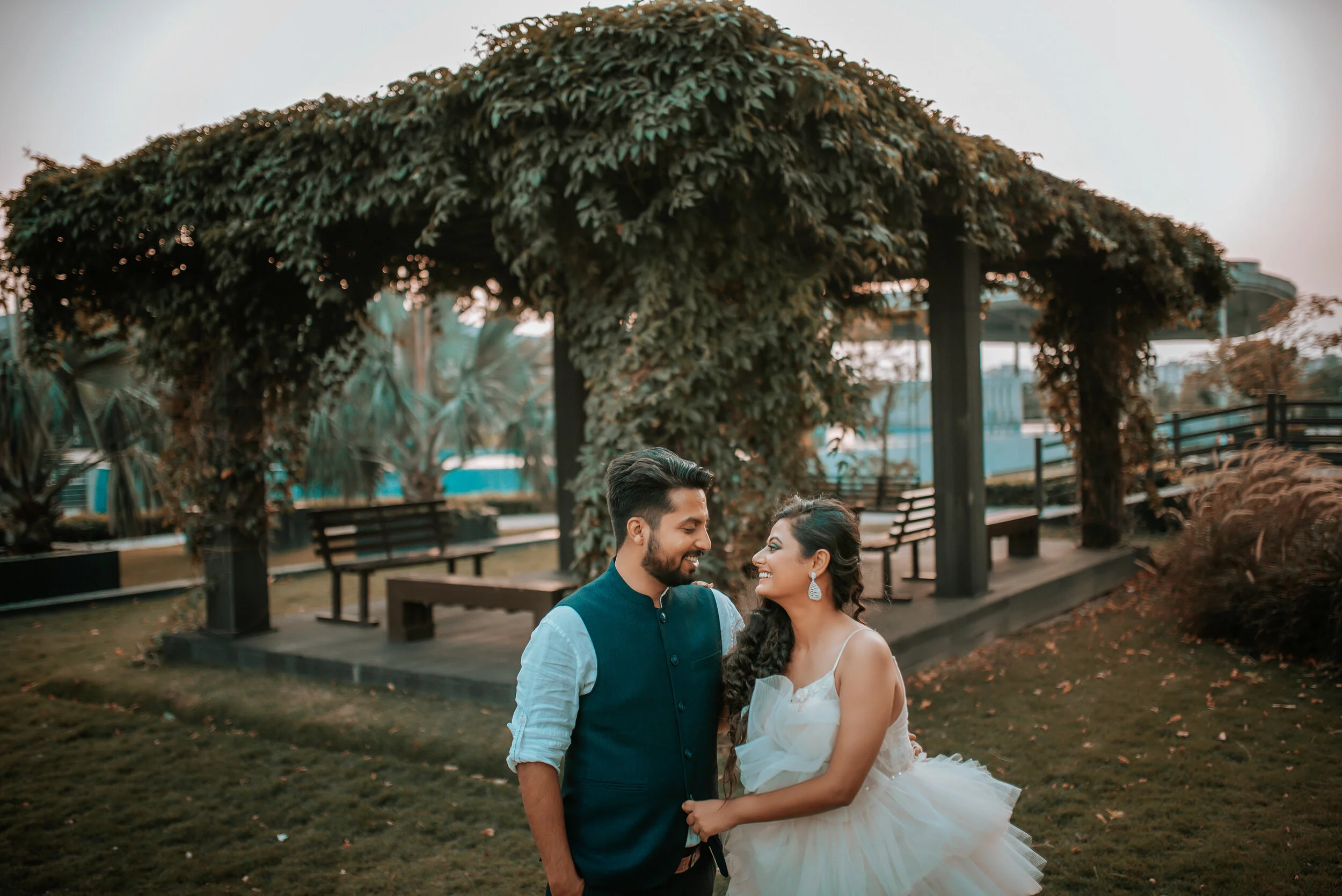 A couple dressed in formal wedding attire standing close to each other in a garden park with a pergola covered in lush greenery behind them, smiling and looking into each other's eyes.