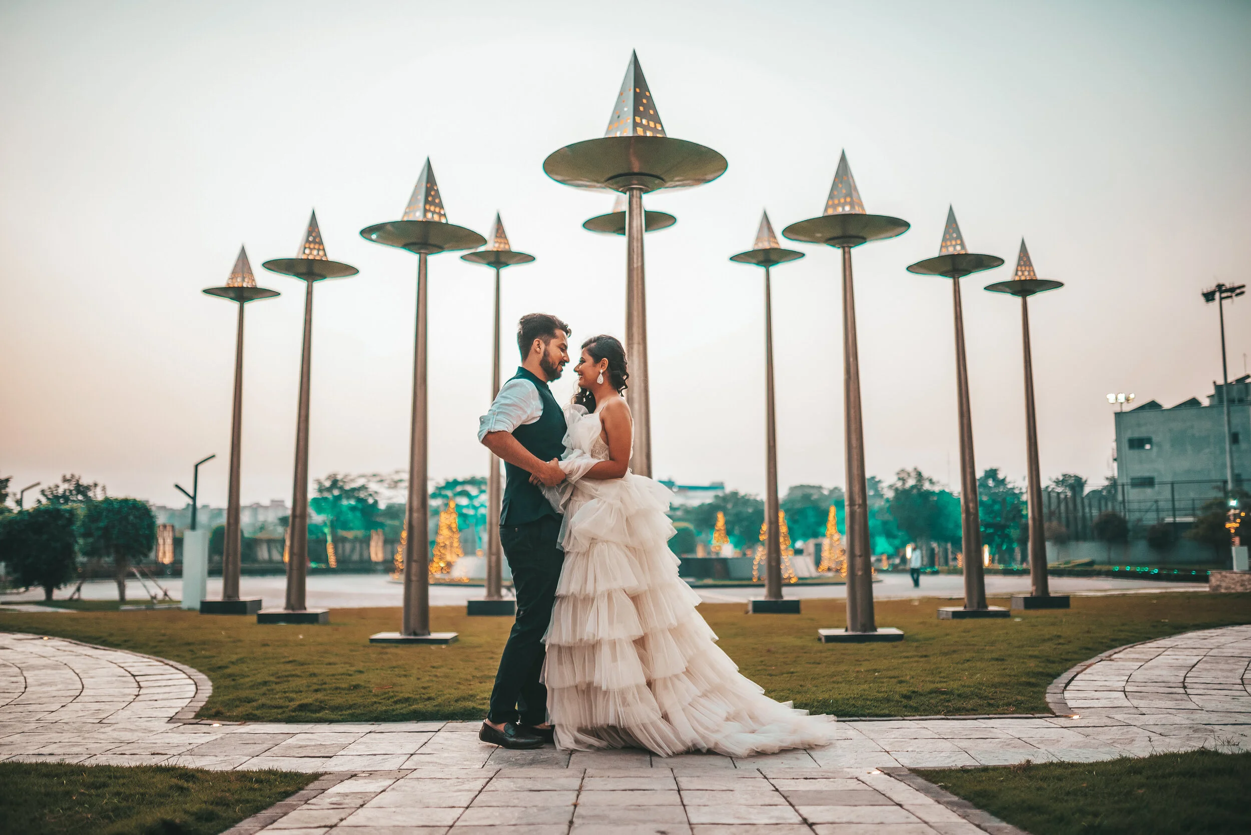 A couple in wedding attire, holding each other closely, standing on a paved walkway outdoors at sunset with decorative lights and umbrella-like structures in the background.