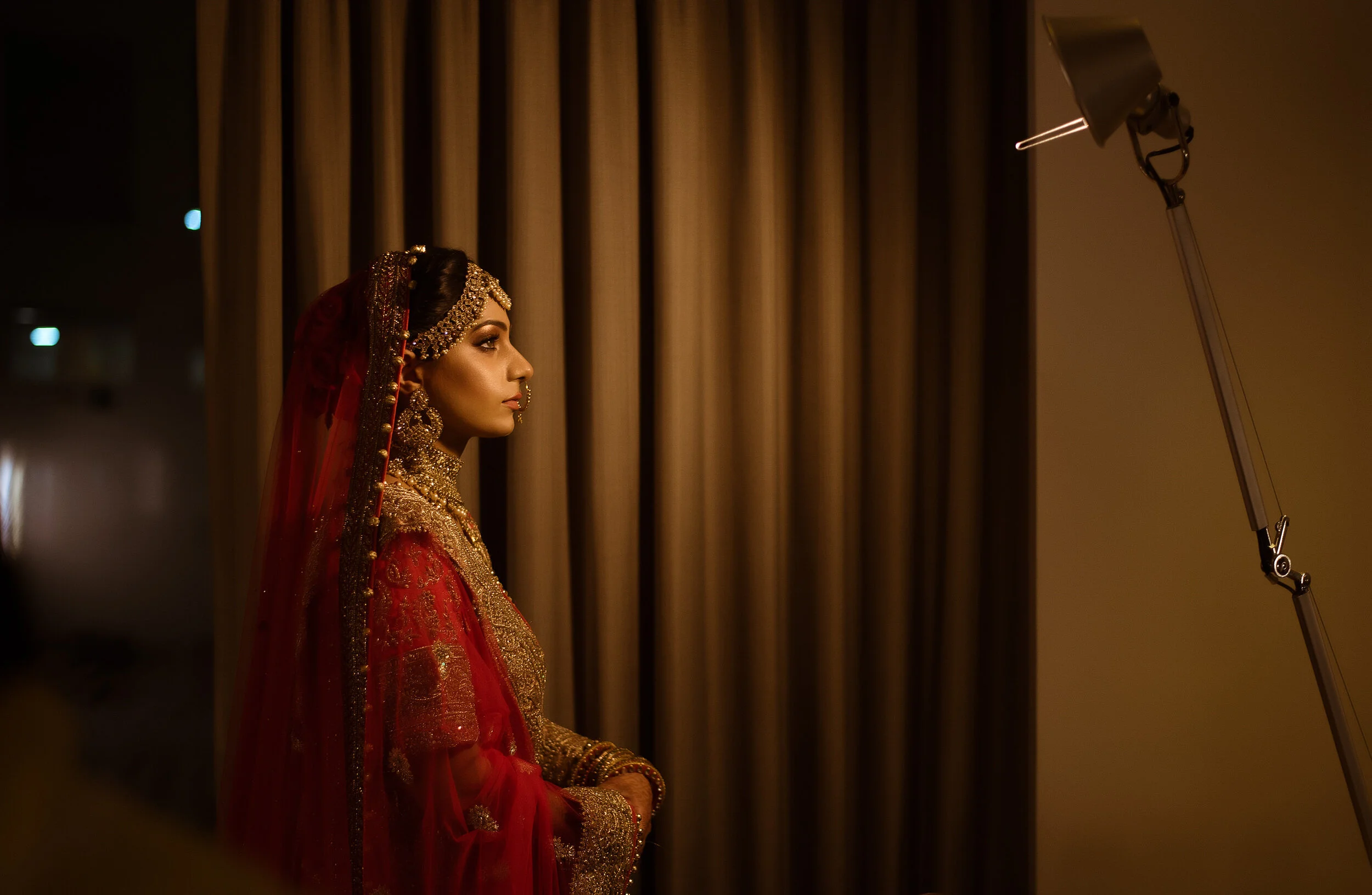 A woman dressed in traditional red and gold bridal attire with jewelry, standing in profile indoors near a curtain, illuminated by a warm light.