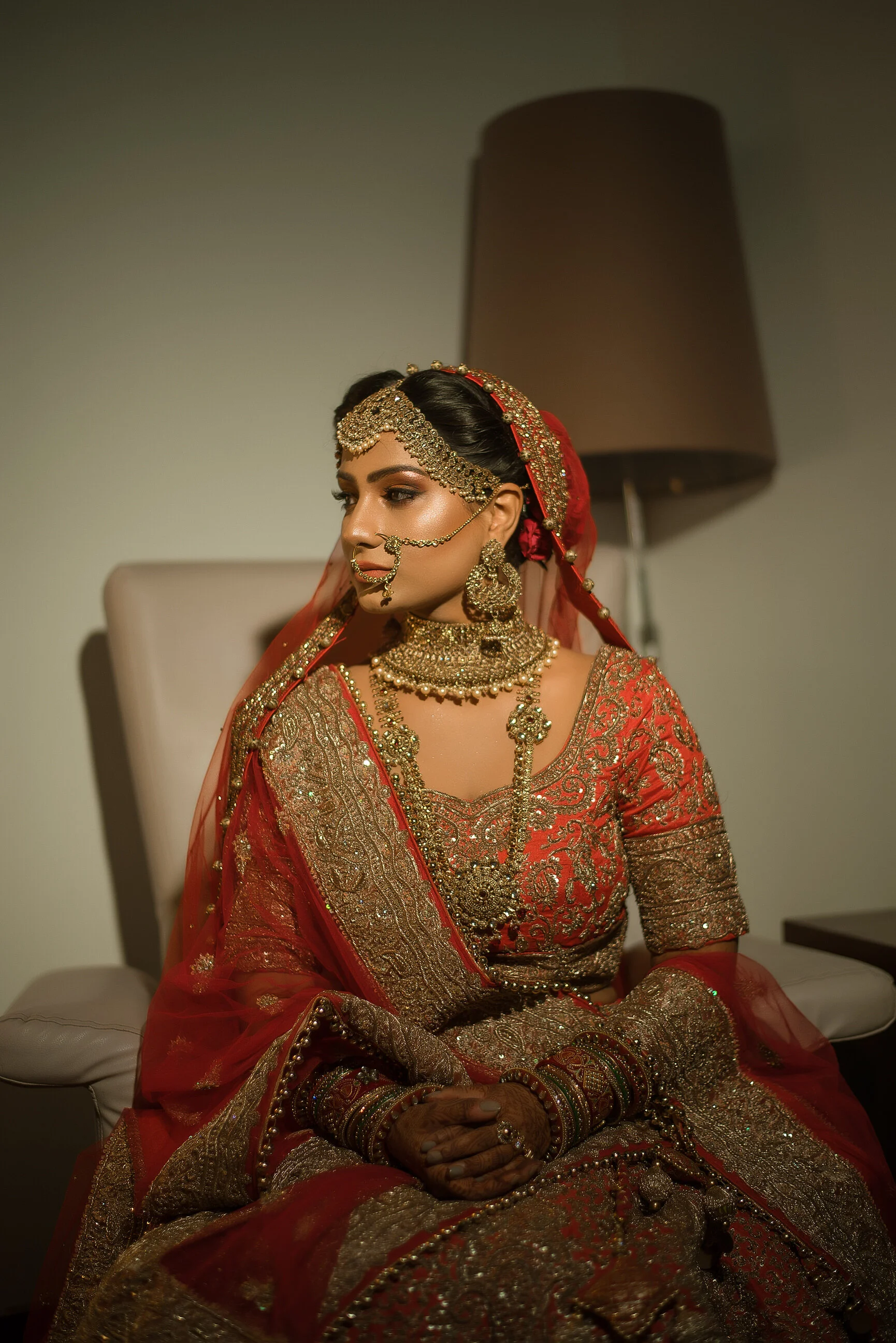 A woman in traditional Indian bridal attire, wearing a richly embroidered red and gold saree, with elaborate gold jewelry, including a maang tikka, nose ring, earrings, necklace, bangles, and henna on her hands, sitting on a chair in an indoor settin