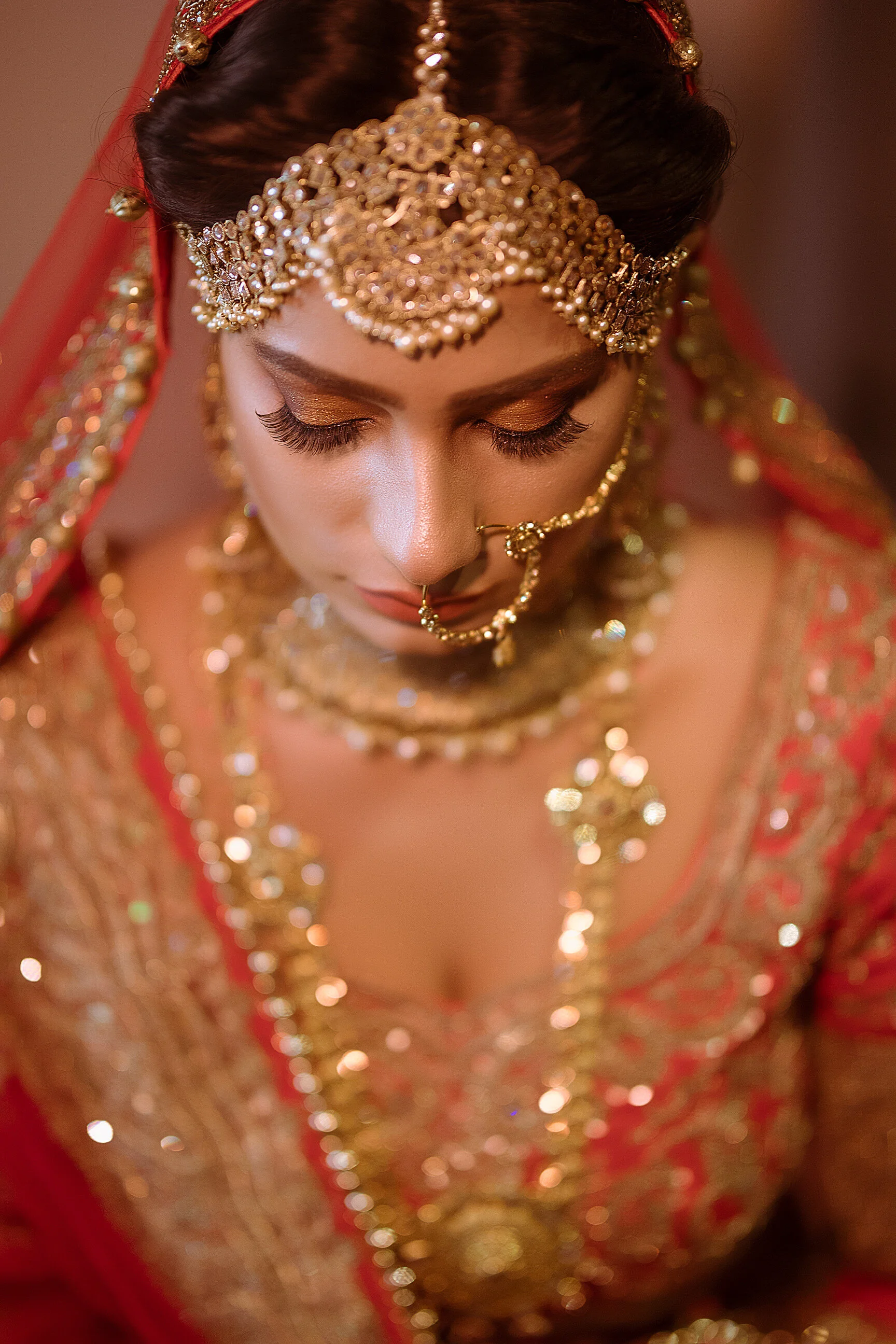 Close-up of a bride wearing elaborate gold jewelry and a red embroidered bridal dress, with her eyes closed and head slightly bowed.