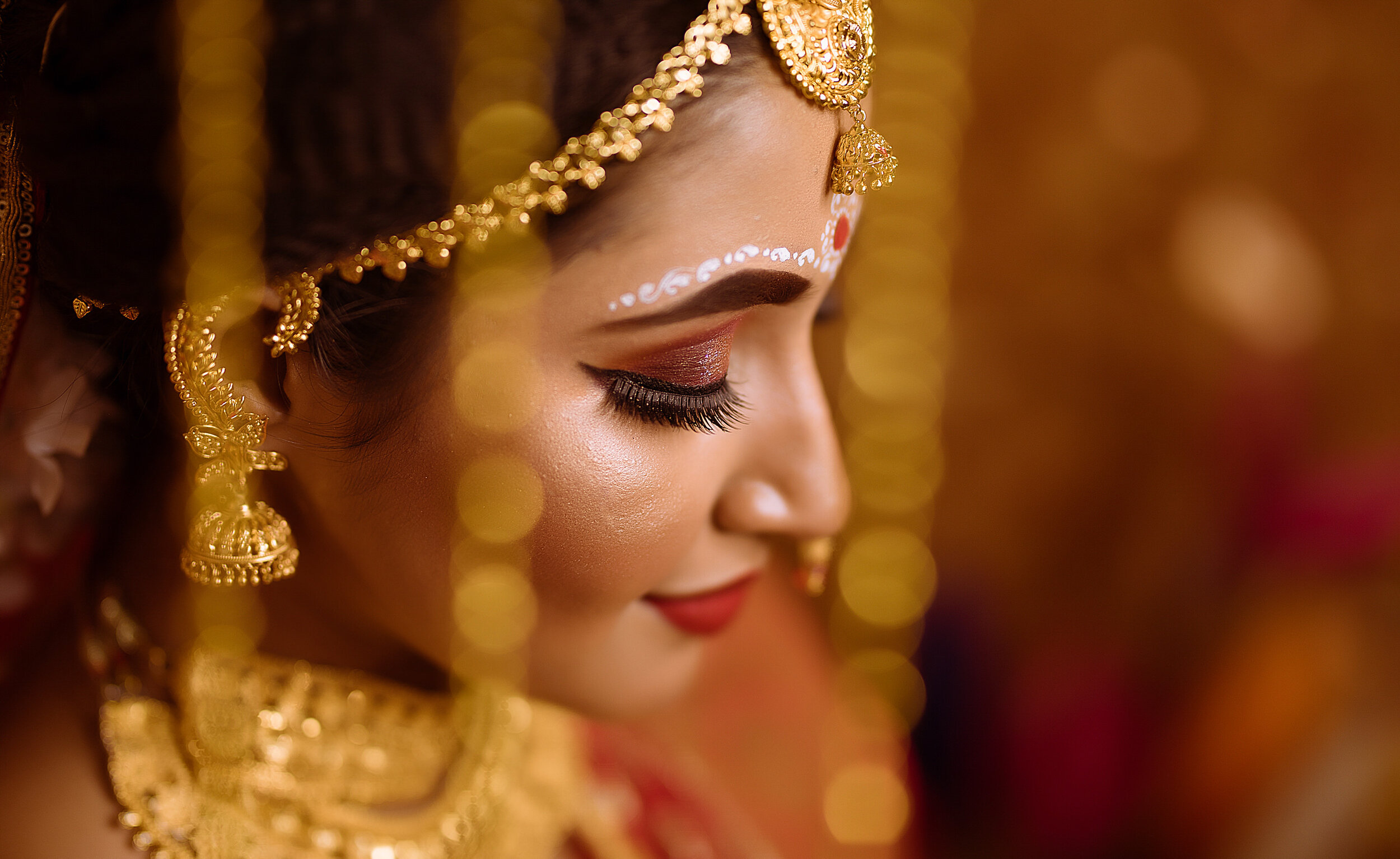 Close-up of an Indian bride wearing traditional gold jewelry and makeup, with her eyes closed, smiling gently.