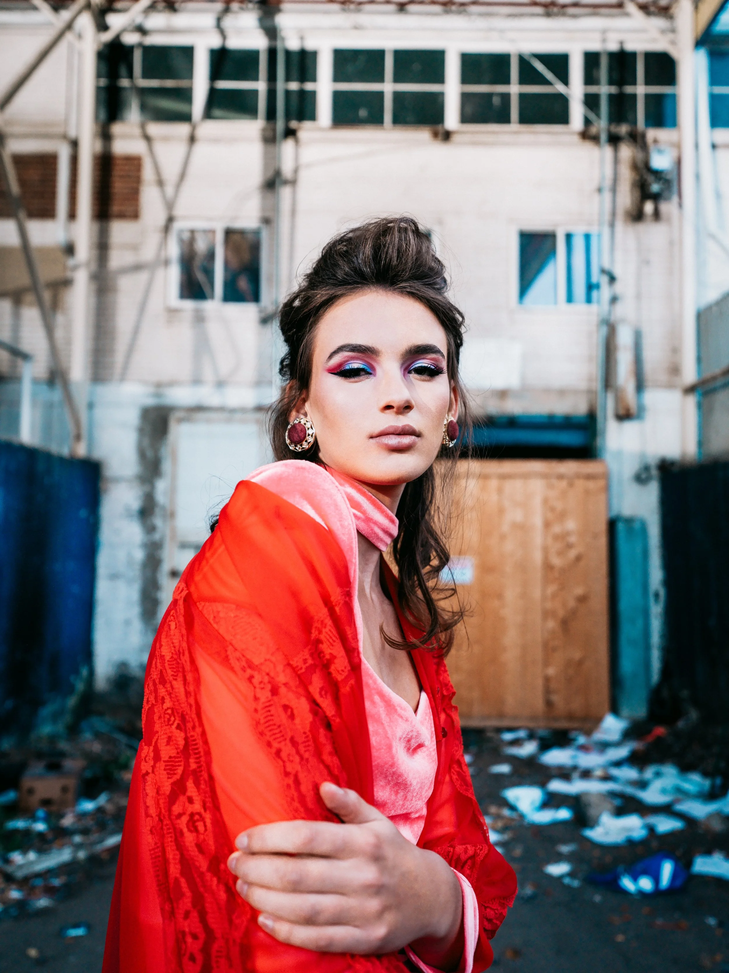 A woman with elaborate makeup and earrings standing outdoors in front of an urban background with trash and a wooden structure.