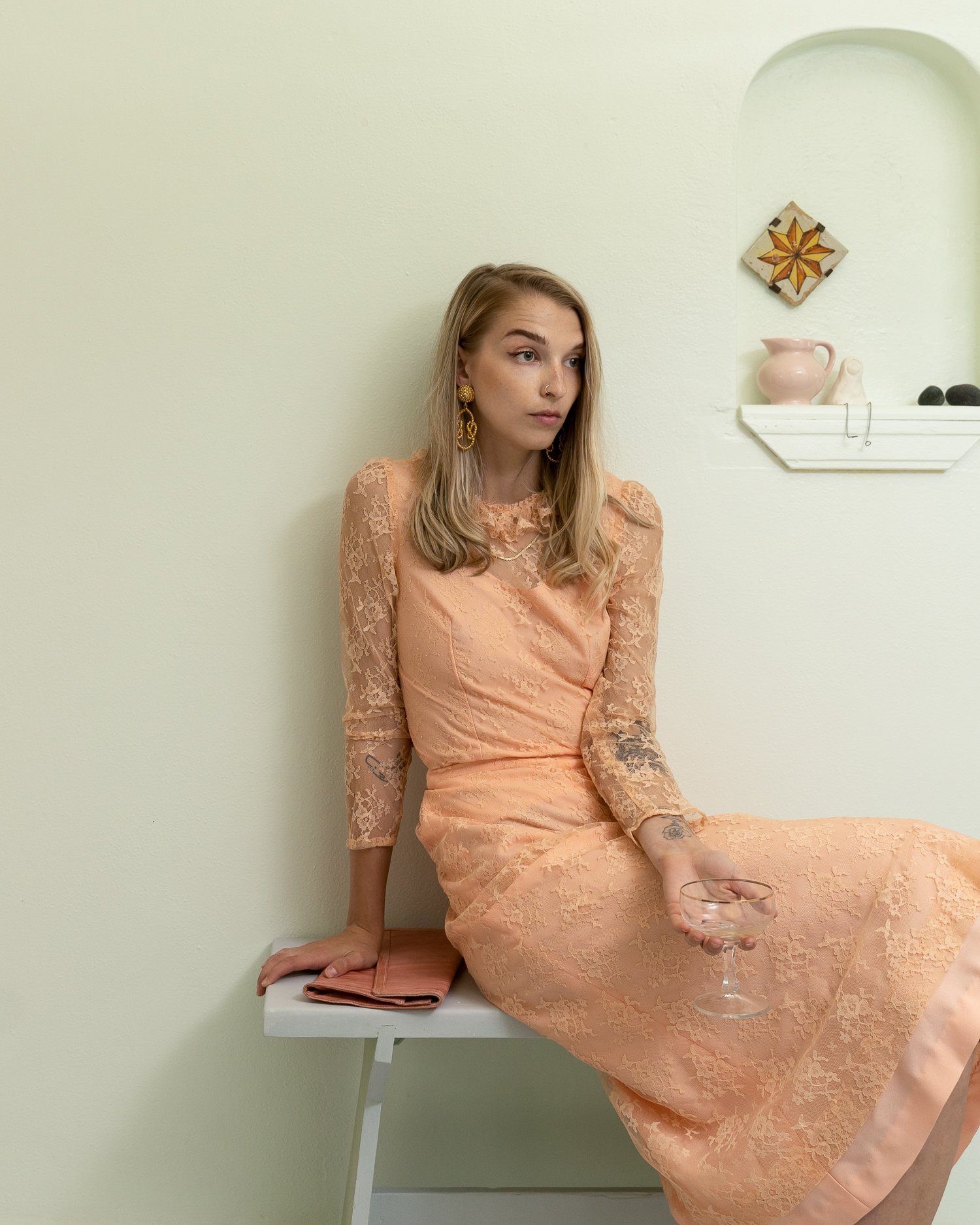 A woman in a peach lace dress sitting on a white bench holding a glass of water, with a small pink cloth and a yellow towel beside her, in a room with light green walls and decorative wall art.