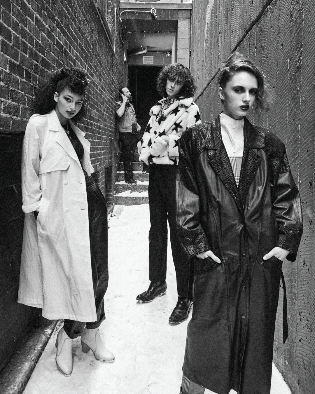 Four women standing in an alleyway with brick and wooden walls. The ground is covered in snow, and one man is in the background talking on a cell phone.