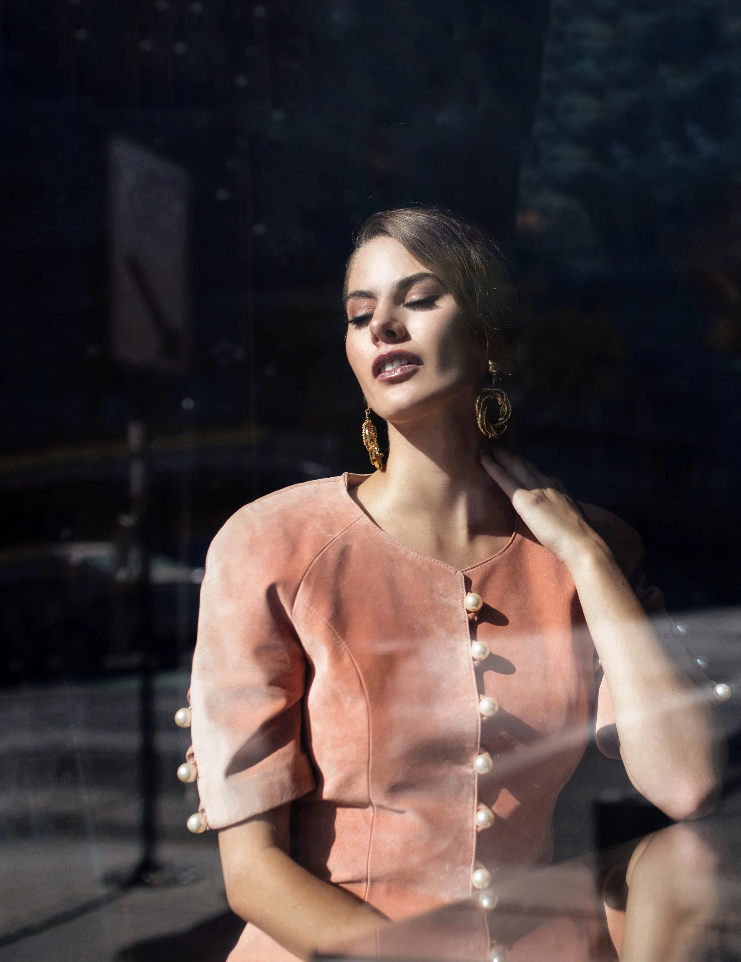 A woman with closed eyes and slightly parted lips, touching her neck, standing behind a glass window. She is wearing a peach-colored dress with pearl button details and large gold earrings. The sunlight highlights her face and upper body.
