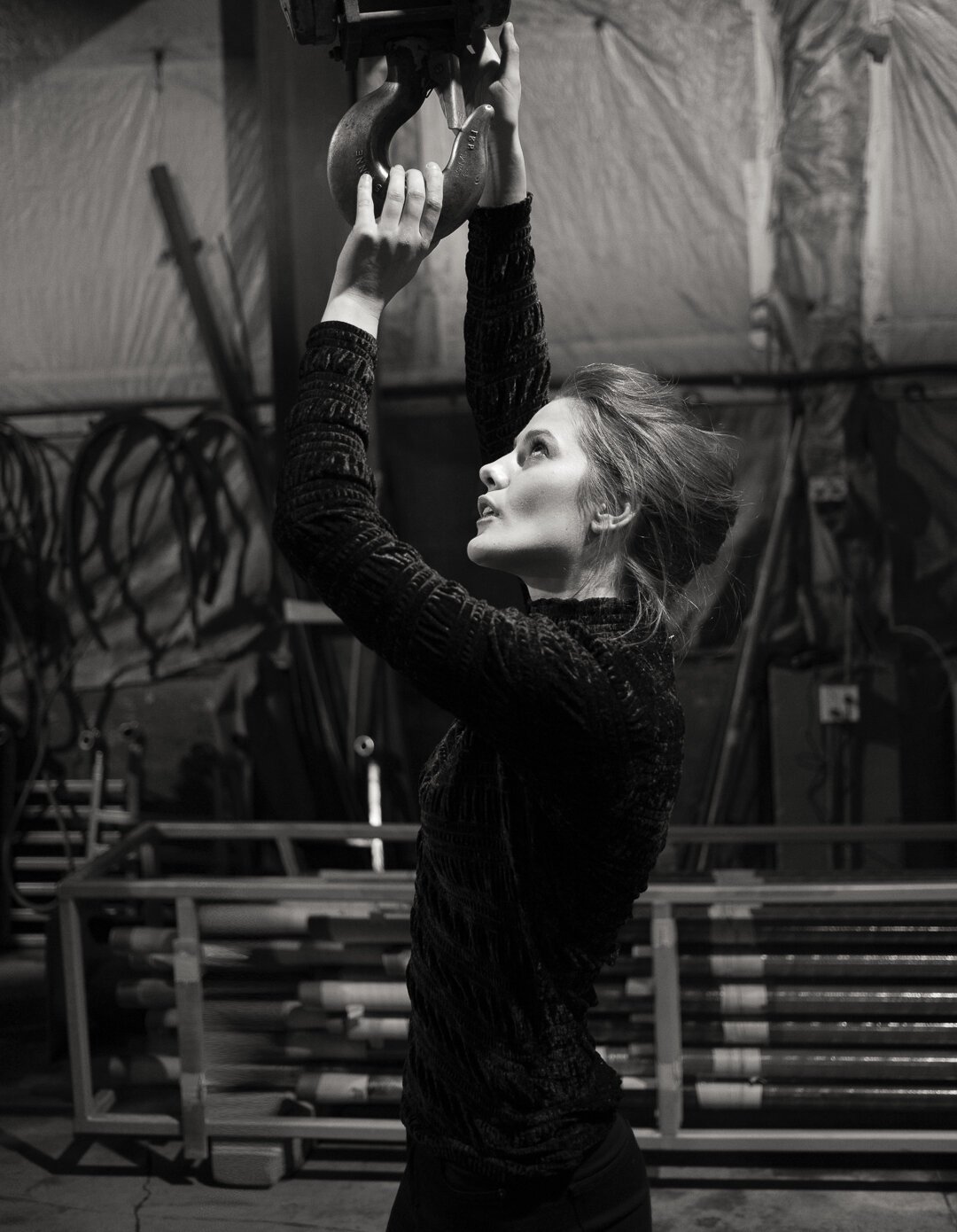 A woman in a black, textured long-sleeve top reaches up to grab a large metal hook hanging from the ceiling in a warehouse or gym setting.