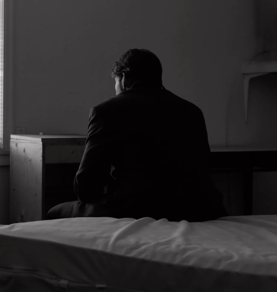 A man wearing a suit sitting on a bed in a dimly lit room, facing away from the camera, looking out a window.