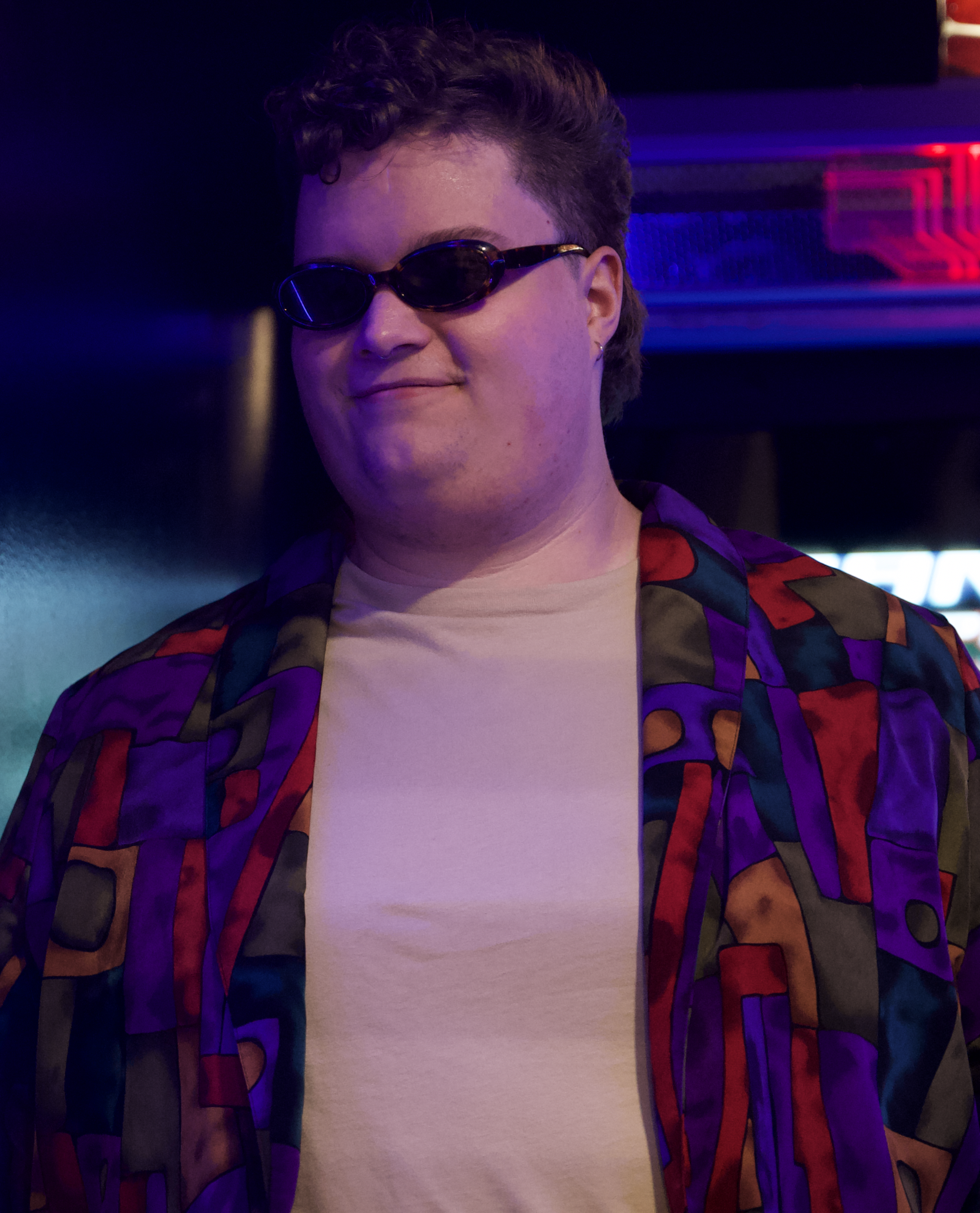 Person with short curly hair wearing sunglasses, a white t-shirt, and a colorful patterned jacket, standing in a dimly lit setting with neon lights in the background.