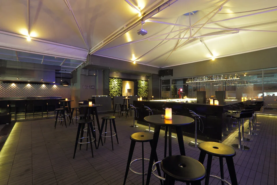 Troika is a spacious bar with a great view, exactly the kind of uncrowded place to hang out in peace. It has a variety of seating arrangements centered around an island bar and is open on all sides, making it a picturesque sight in the evenings. Wooden decking floor, 3 D tiles for wall cladding, all imported lounge furniture and tensile structure for outdoor protection, make it a premium rooftop experience in cool and breezy Bangalore.