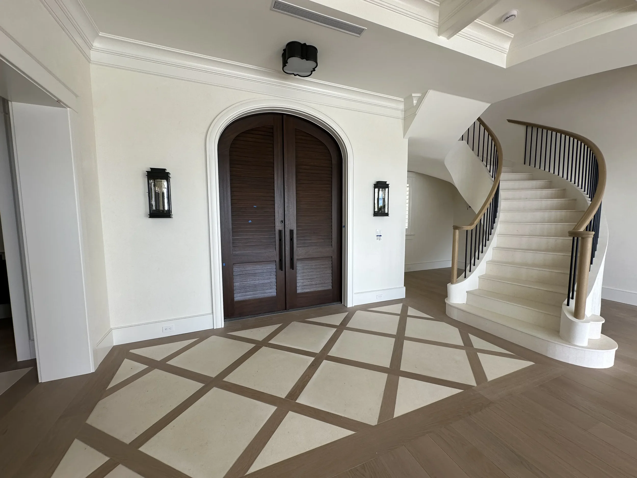 Spacious foyer with arched dark wood double doors, curved staircase, patterned floor inlay, white walls, and wall lanterns.