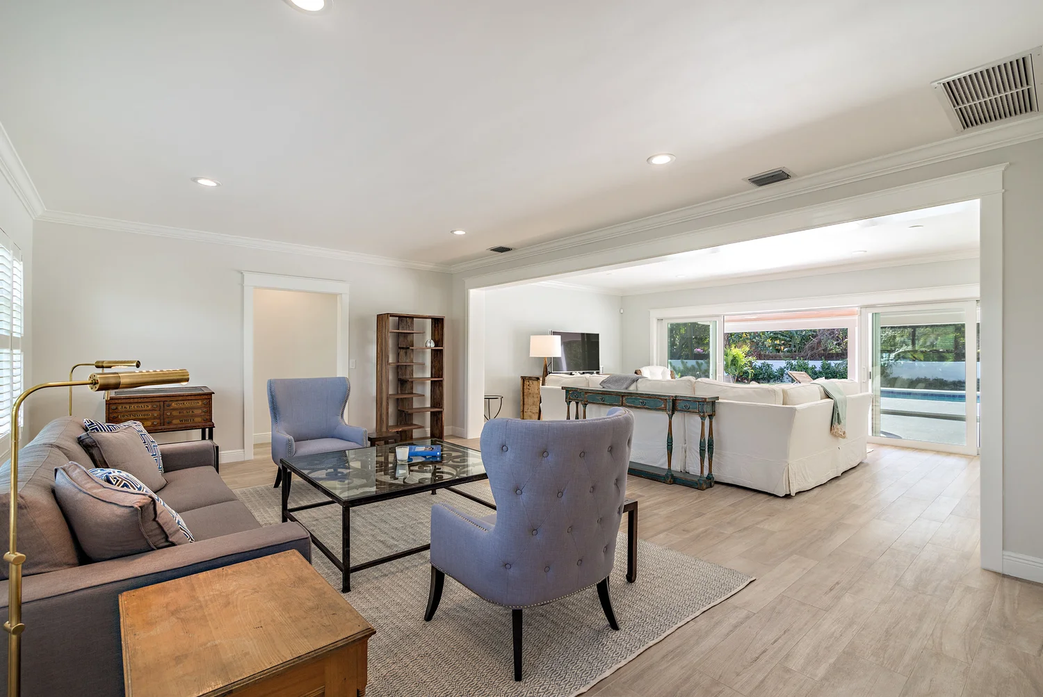 An open concept living room with large windows showing outdoors, featuring a beige sofa with pillows, a glass coffee table, blue armchairs, a wooden side table, a TV with a floor lamp, and a sliding glass door leading outside.