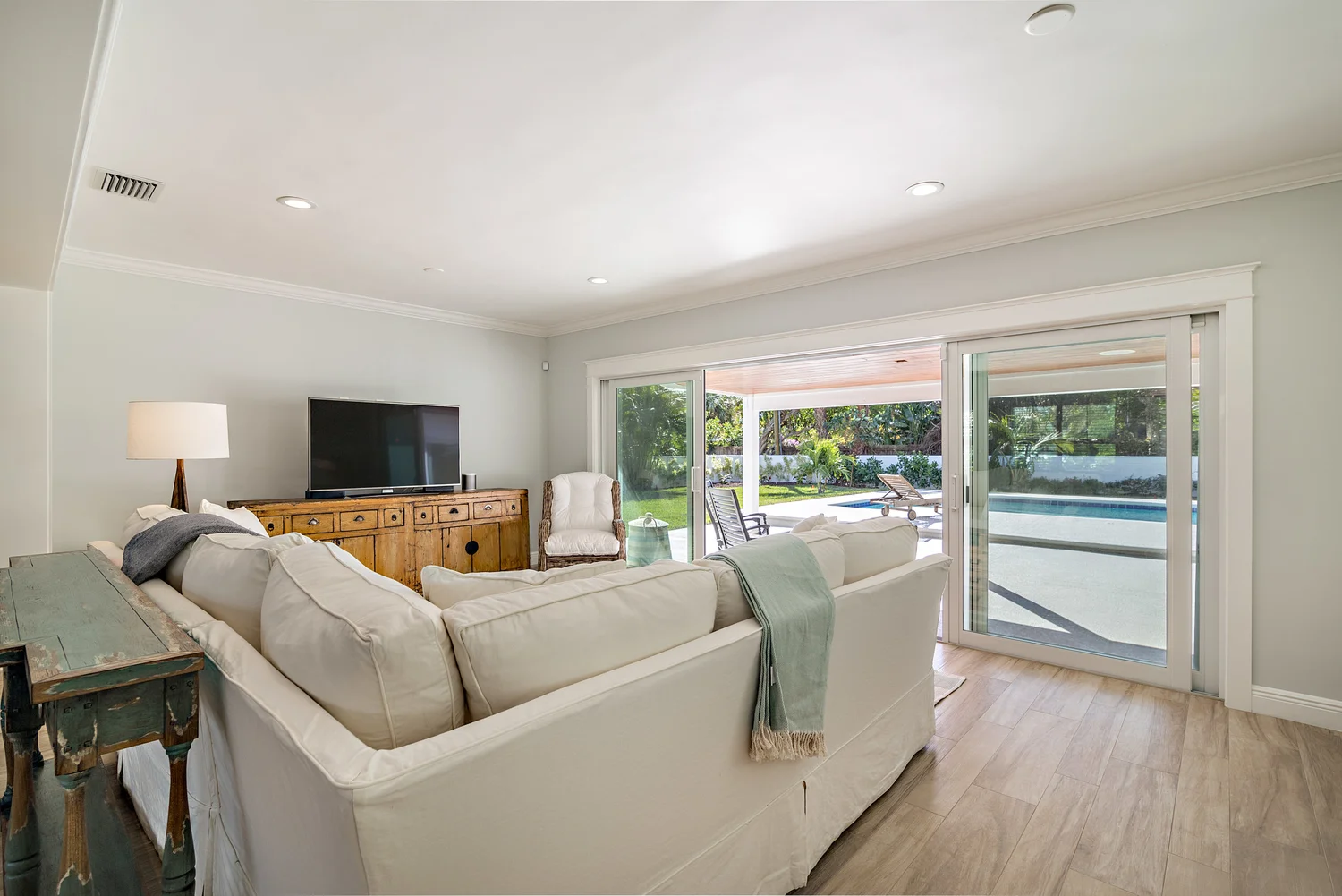 A living room with white sofa, wooden side table, flat-screen TV, and sliding glass doors leading to a backyard with lawn and lounge chairs.