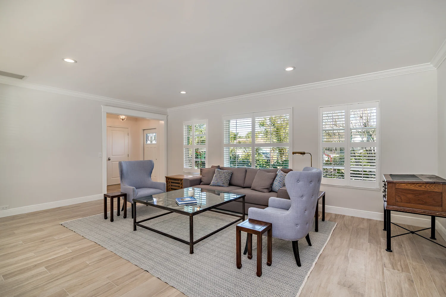 Living room with beige walls, large windows with blinds, gray sofa, armchairs, glass coffee table, wooden side table, and a small rug on light wooden flooring.